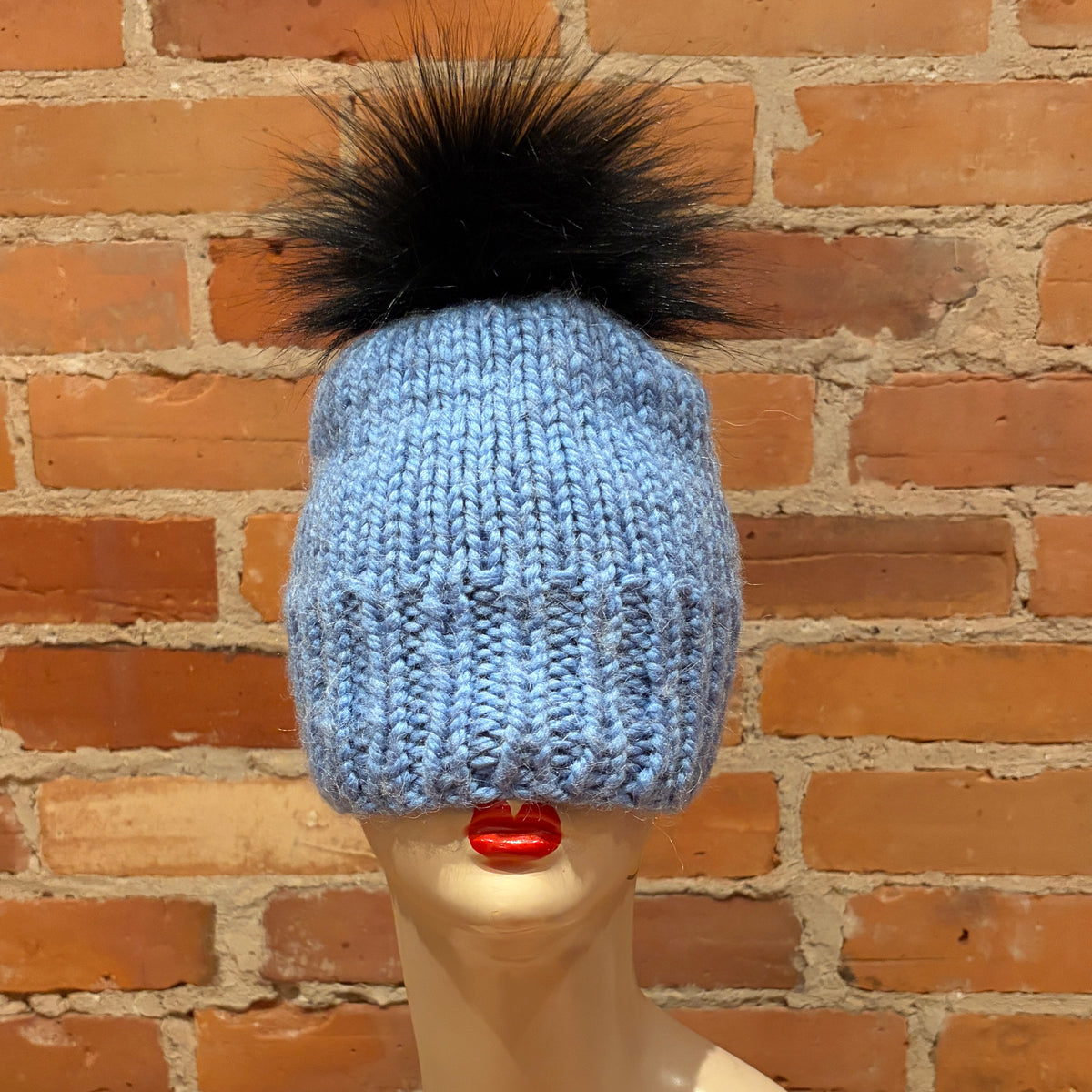 A light blue chunky-knit beanie with a large black faux fur pom-pom, displayed on a mannequin head with red lips in front of a brick wall background.