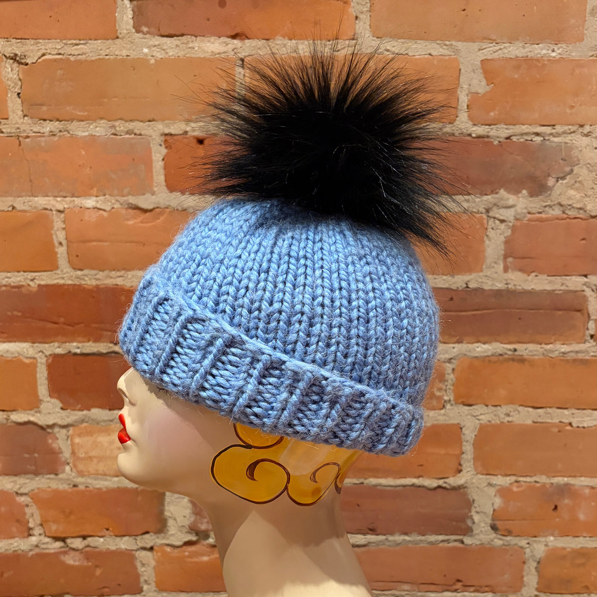 A light blue knit beanie with a large black faux-fur pom-pom, displayed on a mannequin head against a red brick wall background.