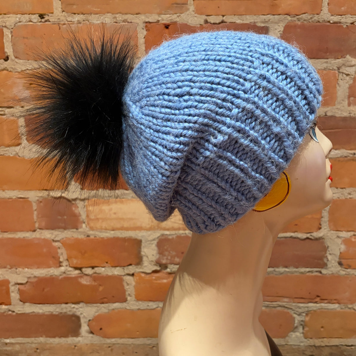 A light blue chunky knit slouchy beanie with a large black faux-fur pom-pom, displayed on a mannequin head against a red brick wall background.