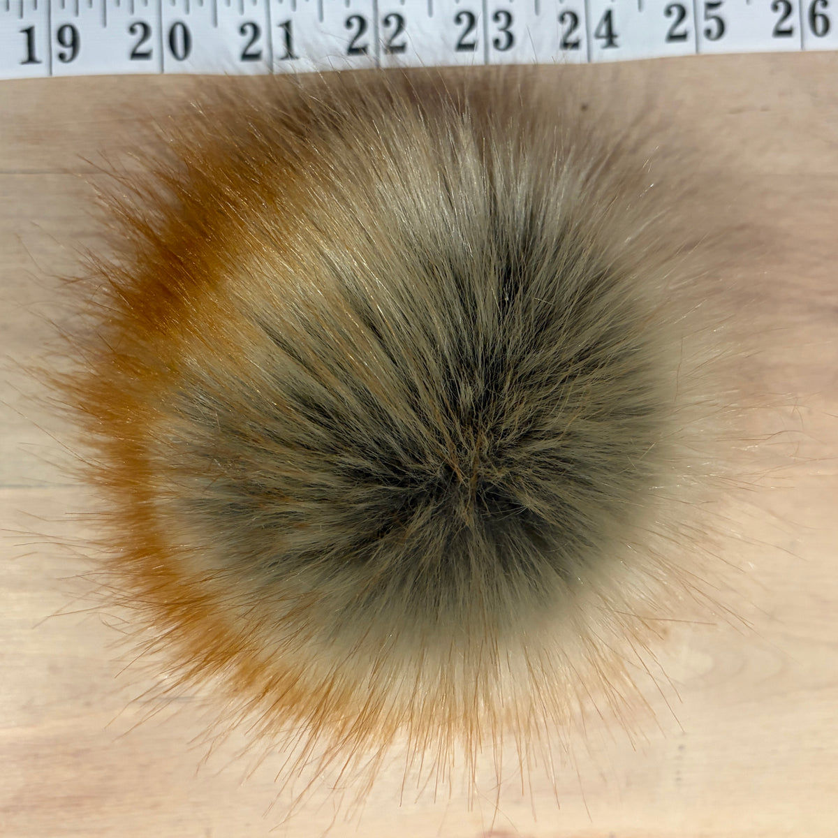 A round faux fur pom-pom with a mix of brown, gray, and beige tones, shown on a light wood surface with a measuring tape visible at the top edge.