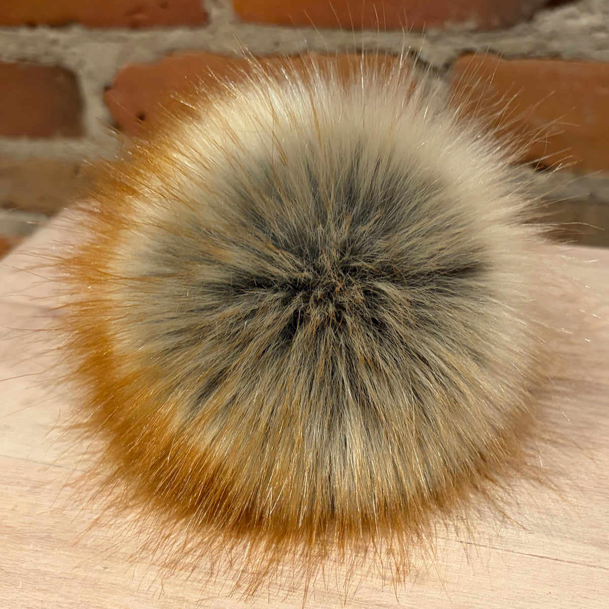 A close-up of a fluffy faux fur pom-pom accessory with a gradient of cream, gray, and golden-brown, resting on a light wood surface with a blurred brick wall background.