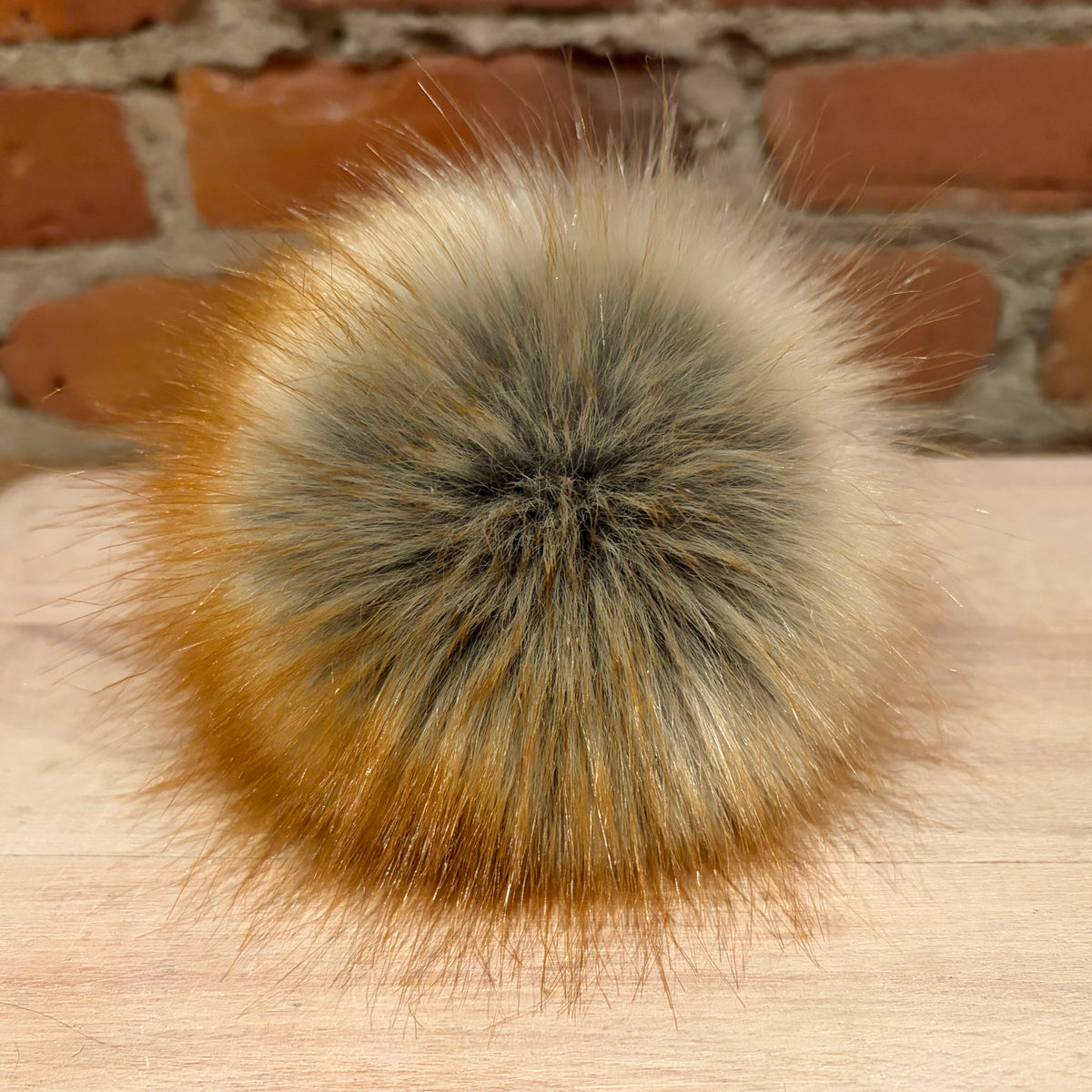 A fluffy faux-fur pom-pom accessory with a gradient of cream, gray, and warm brown, resting on a light wood surface with a brick wall background.