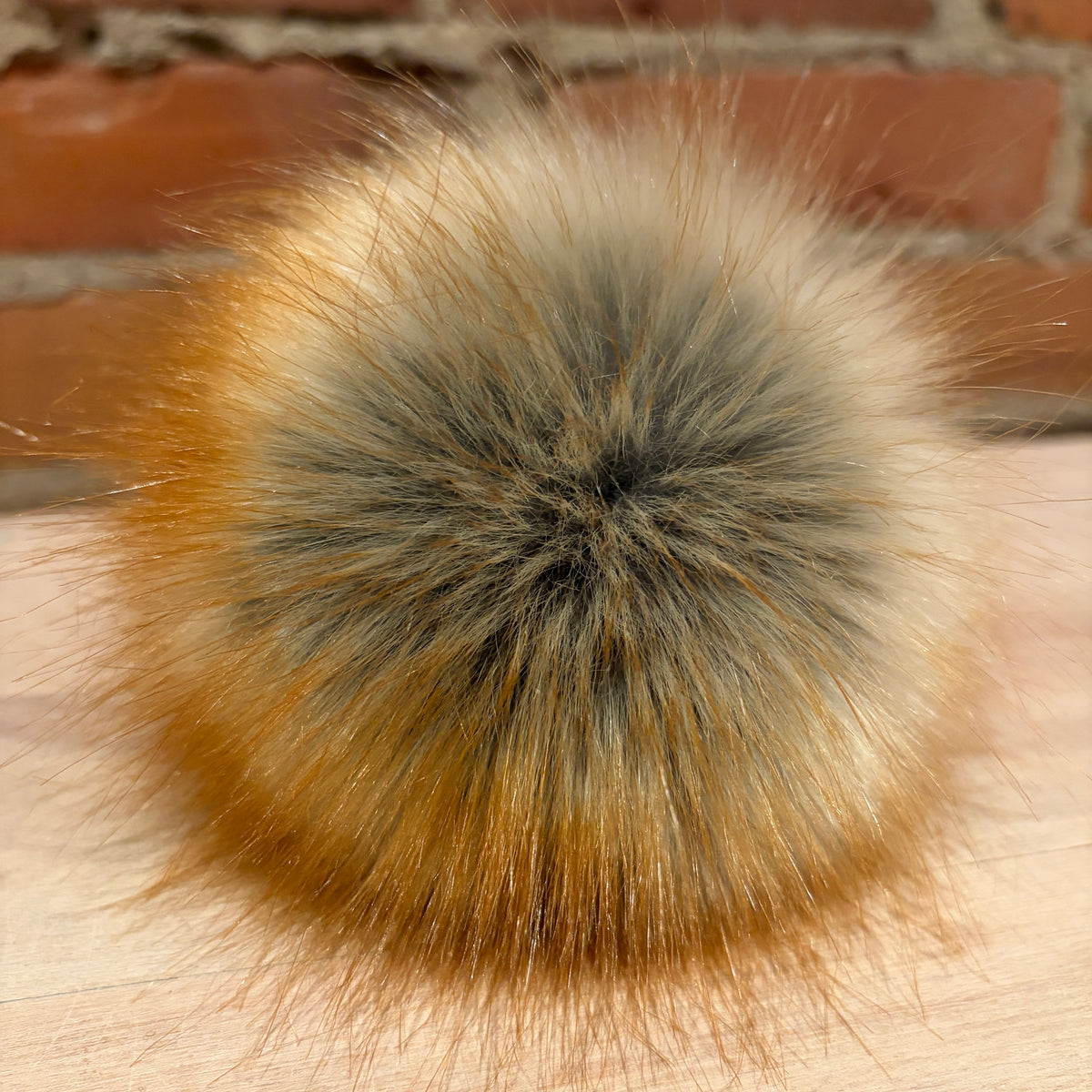 A close-up of a fluffy faux-fur pom pom with a gradient of orange, beige, and gray, resting on a light wood surface with a brick wall background.