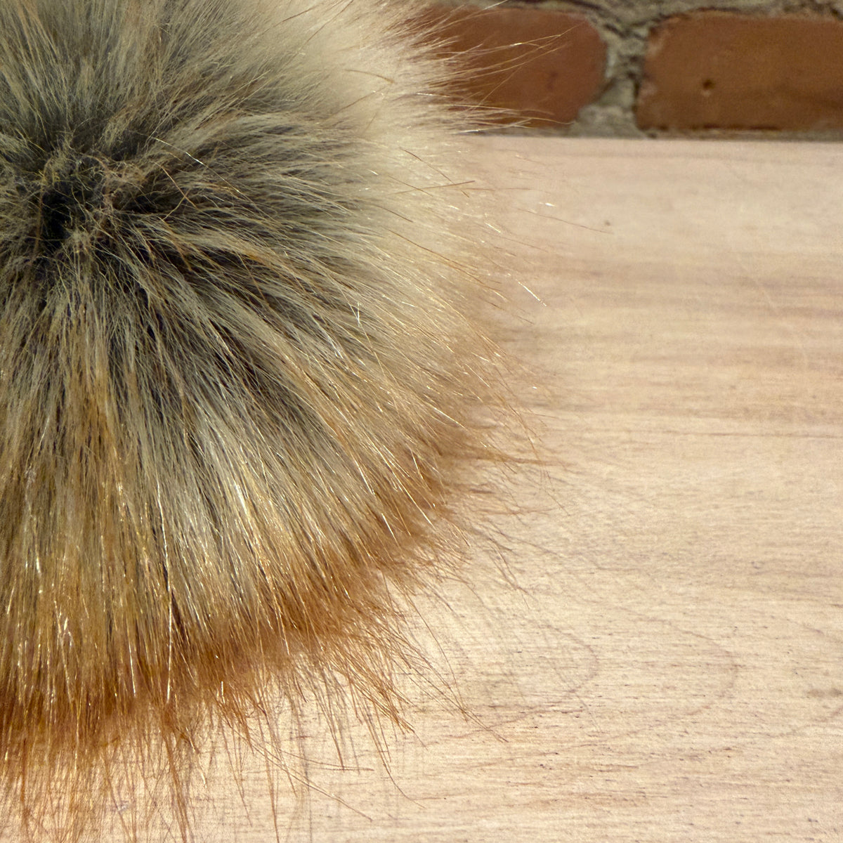 A close-up of a fluffy faux fur pom-pom in shades of beige, brown, and gray, resting on a light wood surface with a brick wall in the background.
