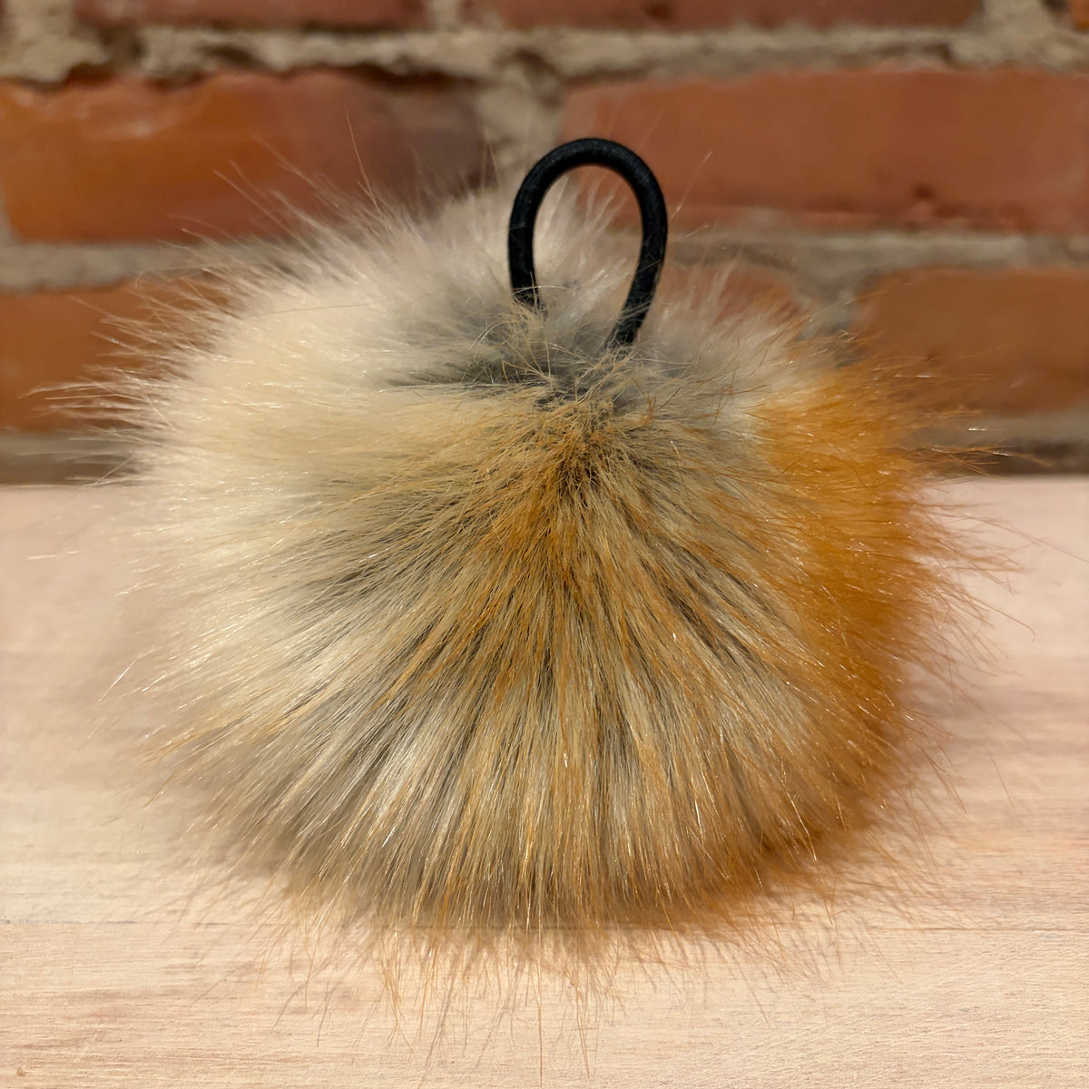 A fluffy faux-fur pom-pom keychain or bag charm with a black plastic loop, featuring a blend of cream, tan, and golden-brown tones, resting on a light wood surface with a brick wall background.