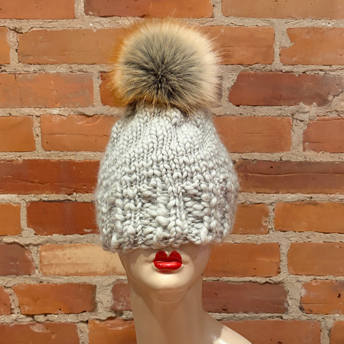 A chunky knit beanie hat in light gray with a large fluffy pom-pom in beige, brown, and gray, displayed on a mannequin head with red lips in front of a red brick wall.