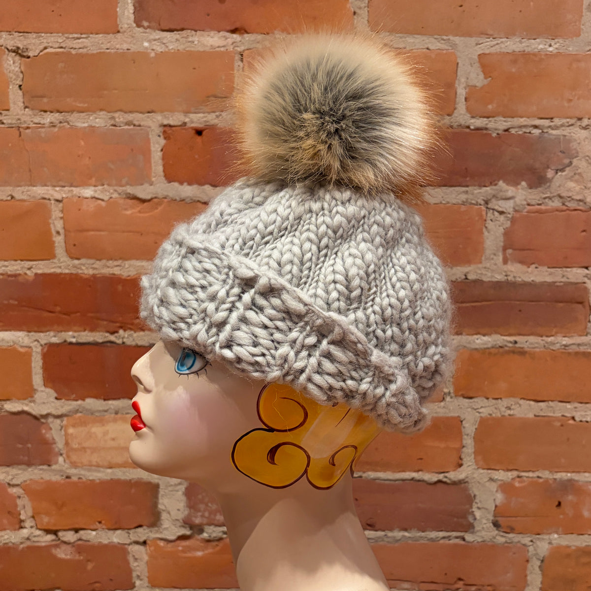 A chunky knit winter hat in light gray with a large beige and brown faux fur pom-pom, displayed on a mannequin head against a red brick wall background.