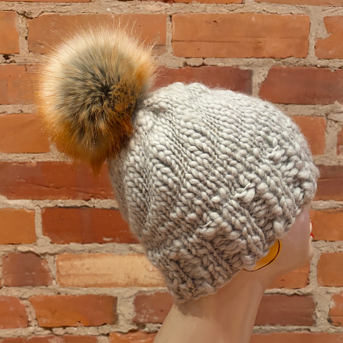 A chunky knit beanie hat in a light heather gray color, featuring a large fluffy faux-fur pom-pom in shades of brown, beige, and gray. The hat is displayed on a mannequin head against a rustic red brick wall background.