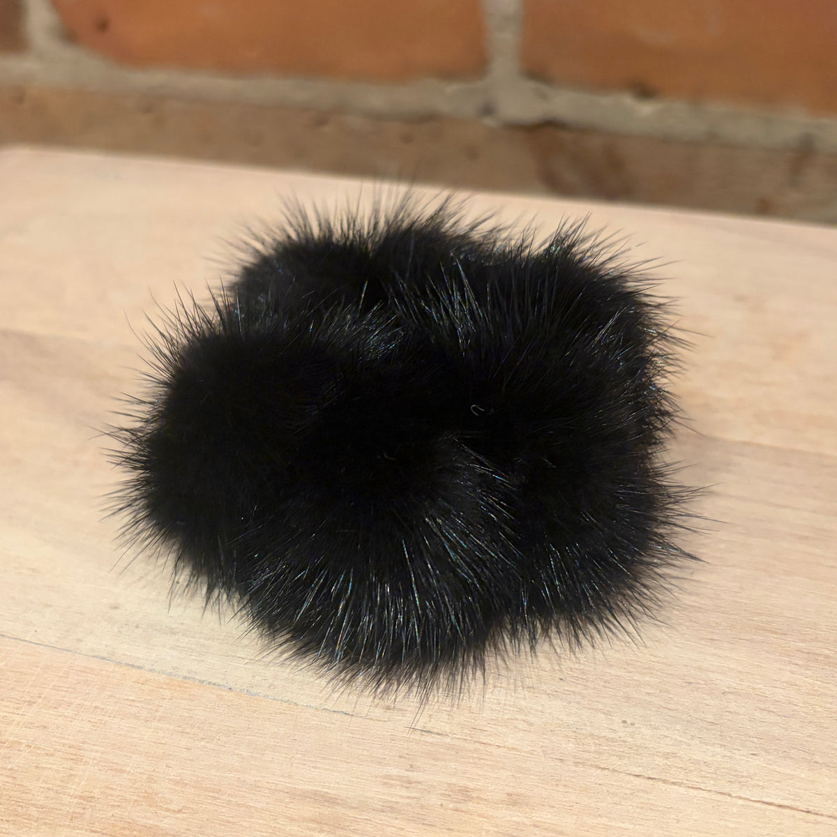 A black mink fur pom-pom scrunchie resting on a light-colored wooden surface with a blurred brick wall in the background.