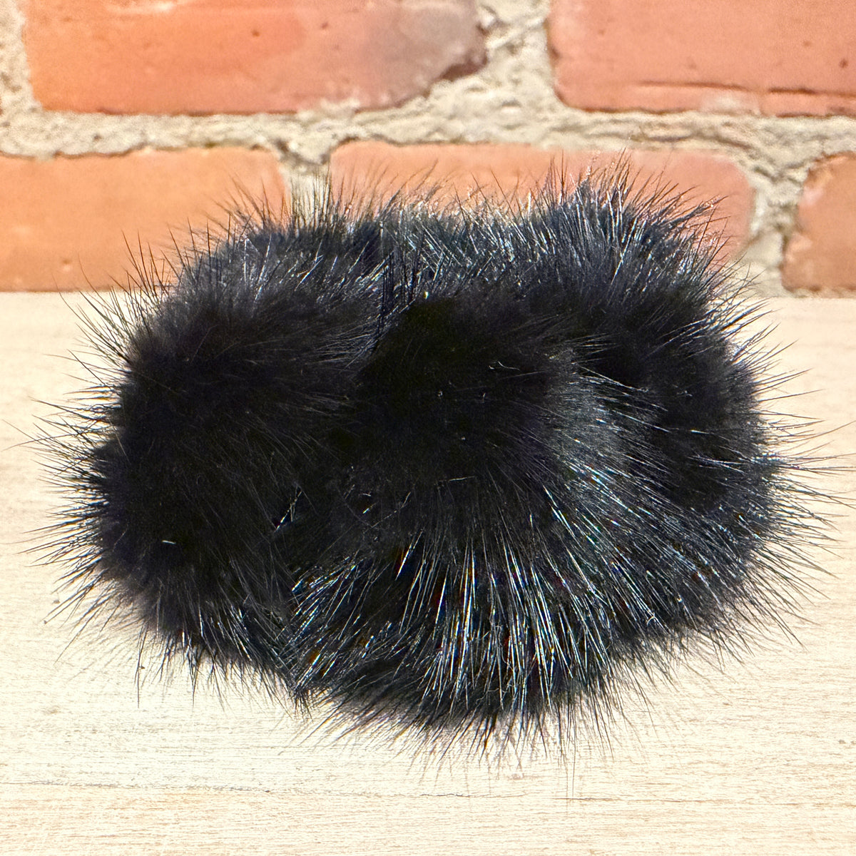 A black faux fur pom-pom accessory, likely a hair claw clip or decorative pom-pom, resting on a light wood surface with a red brick wall background.