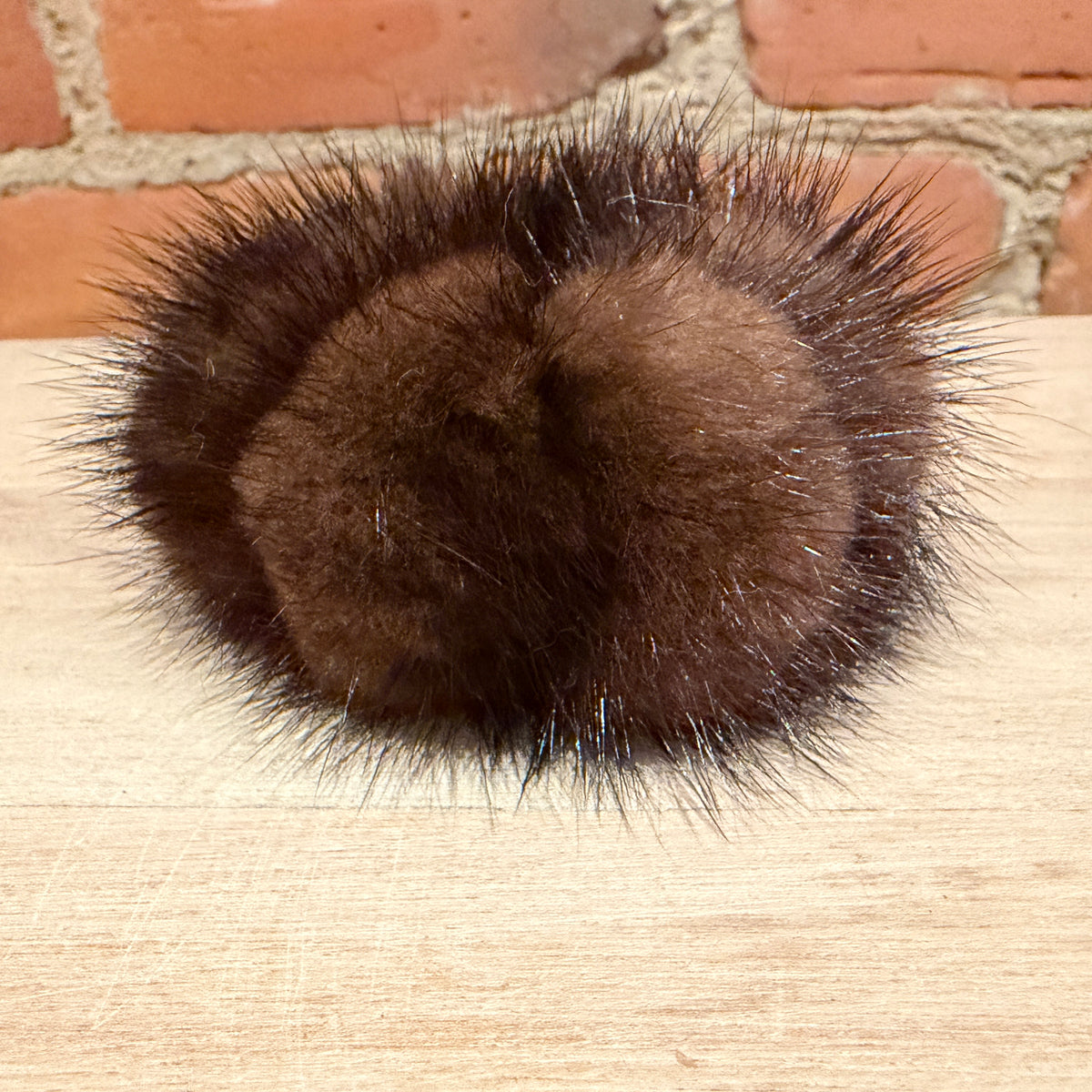 A dark brown fluffy mink fur hair scrunchie, resting on a light wood surface with a brick wall background.