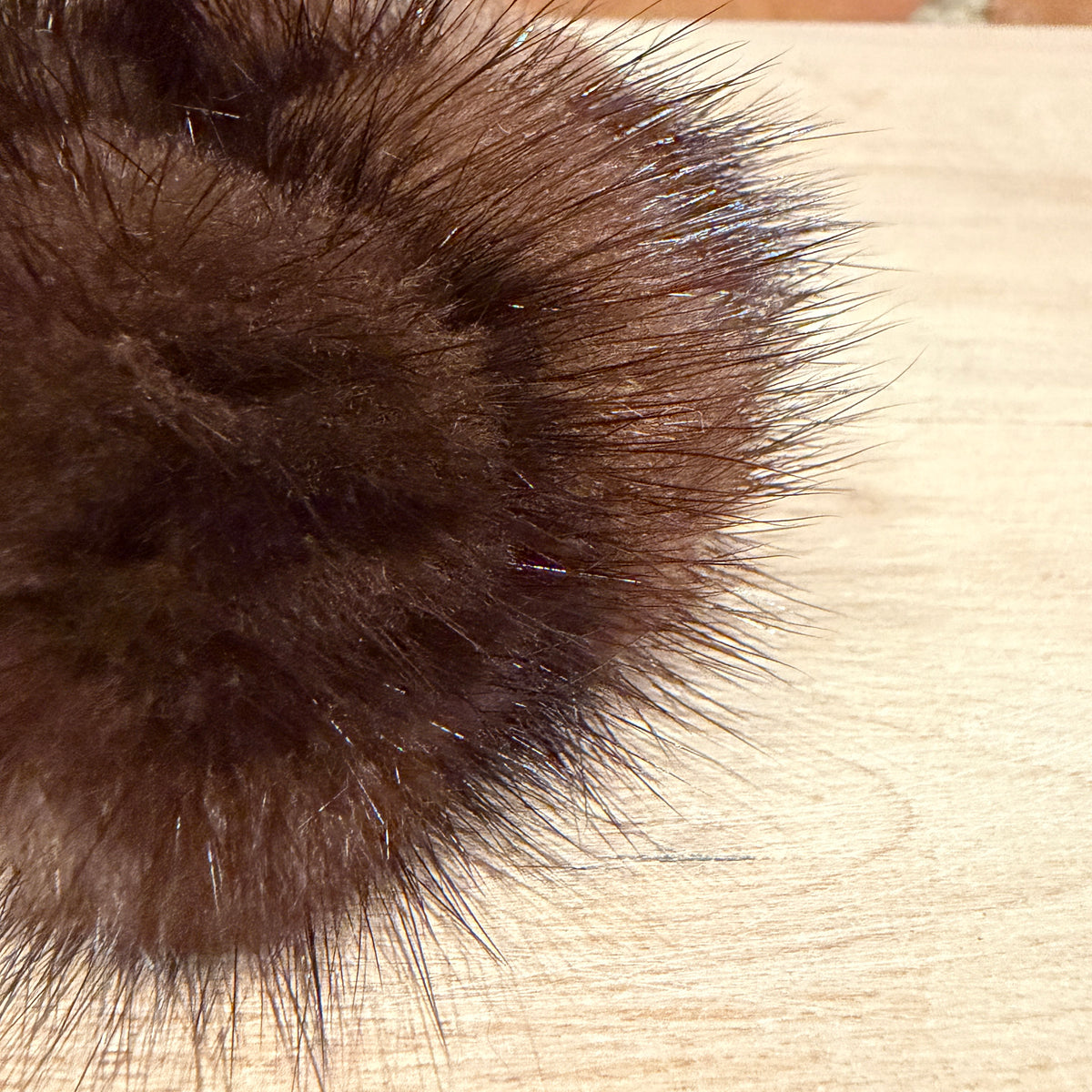 A close-up of a dark brown mink fur pom-pom accessory resting on a light wood surface.