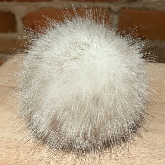 A fluffy white faux fur pom-pom ball resting on a light wood surface with a blurred brick wall background.