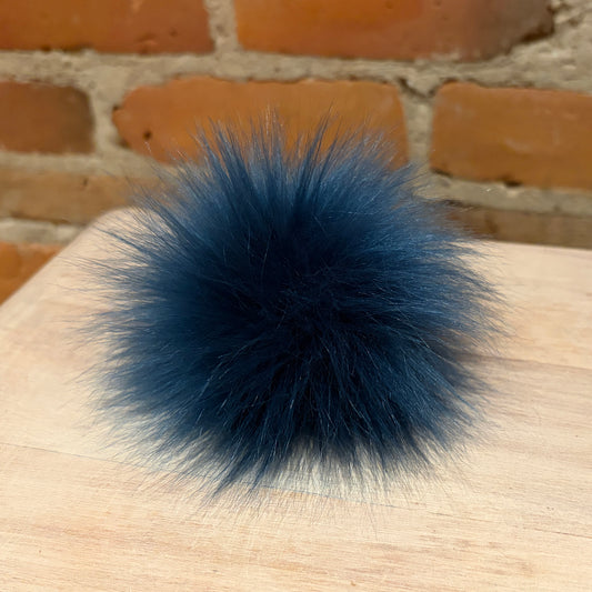 A fluffy dark blue faux-fur pom-pom accessory resting on a light wood surface with a brick wall background.