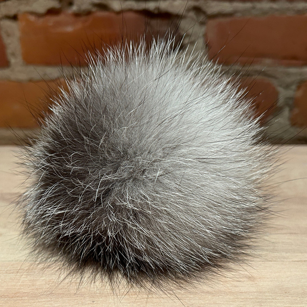 A fluffy gray and black fox fur pom-pom ball resting on a light wood surface with a blurred brick wall background.
