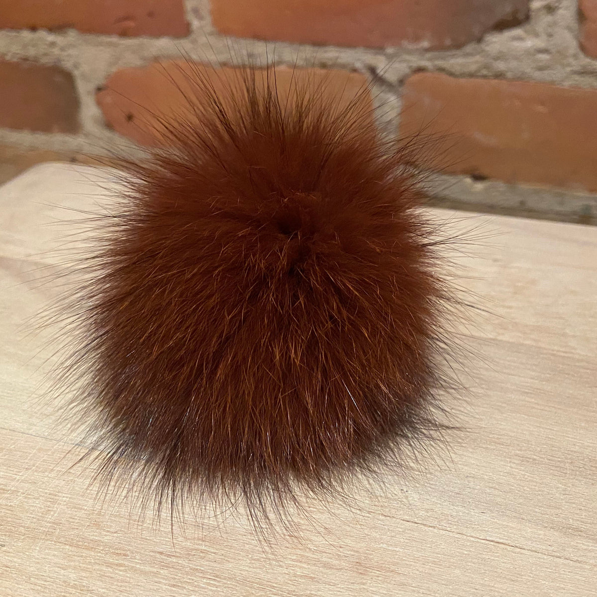 A fluffy, reddish-brown fox fur pom-pom ball resting on a light-colored wooden surface with a brick wall background.