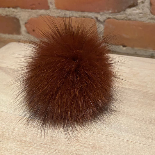 A fluffy, reddish-brown fox fur pom-pom ball resting on a light-colored wooden surface with a brick wall background.