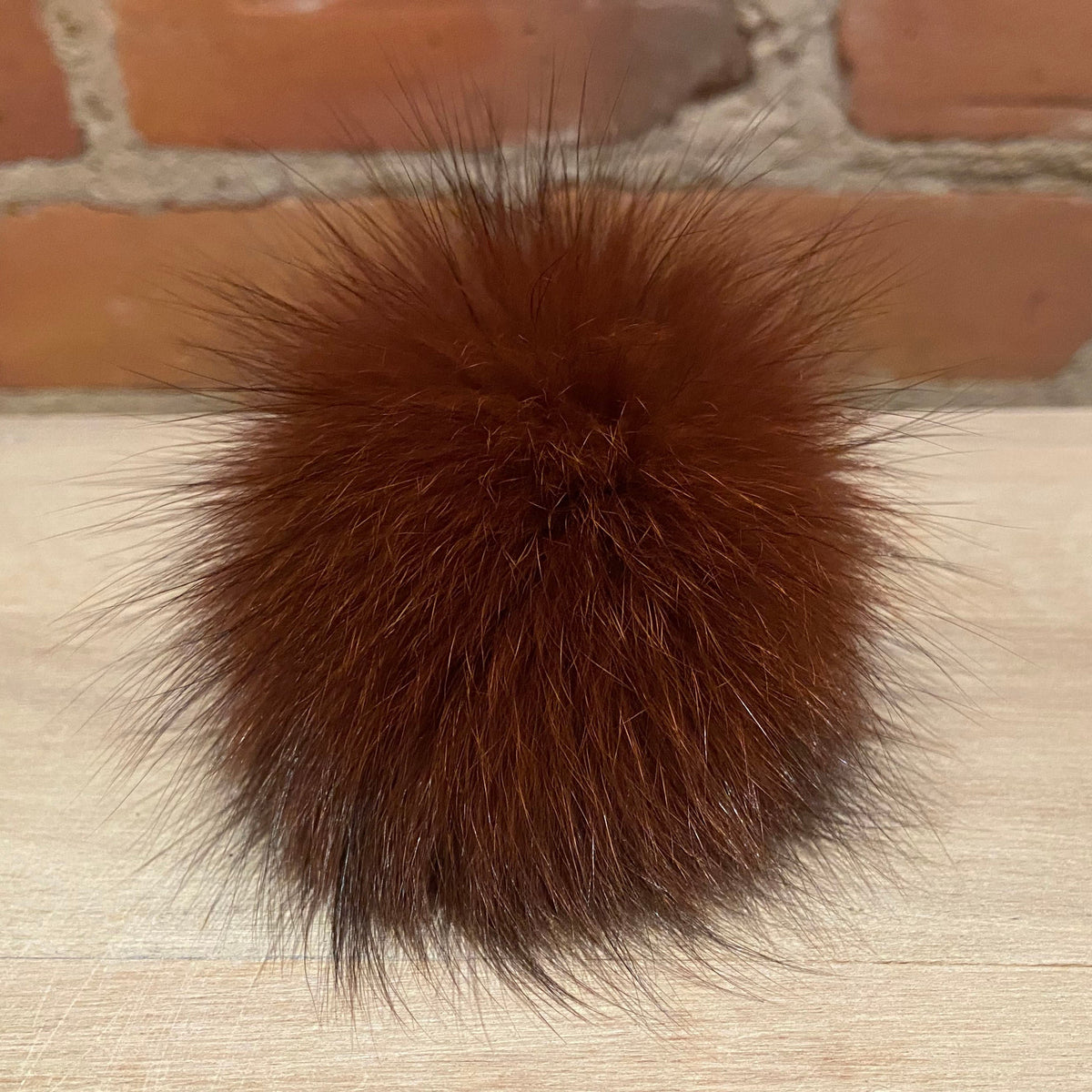 A round, fluffy brown fur pom-pom accessory resting on a light wood surface with a brick wall background.