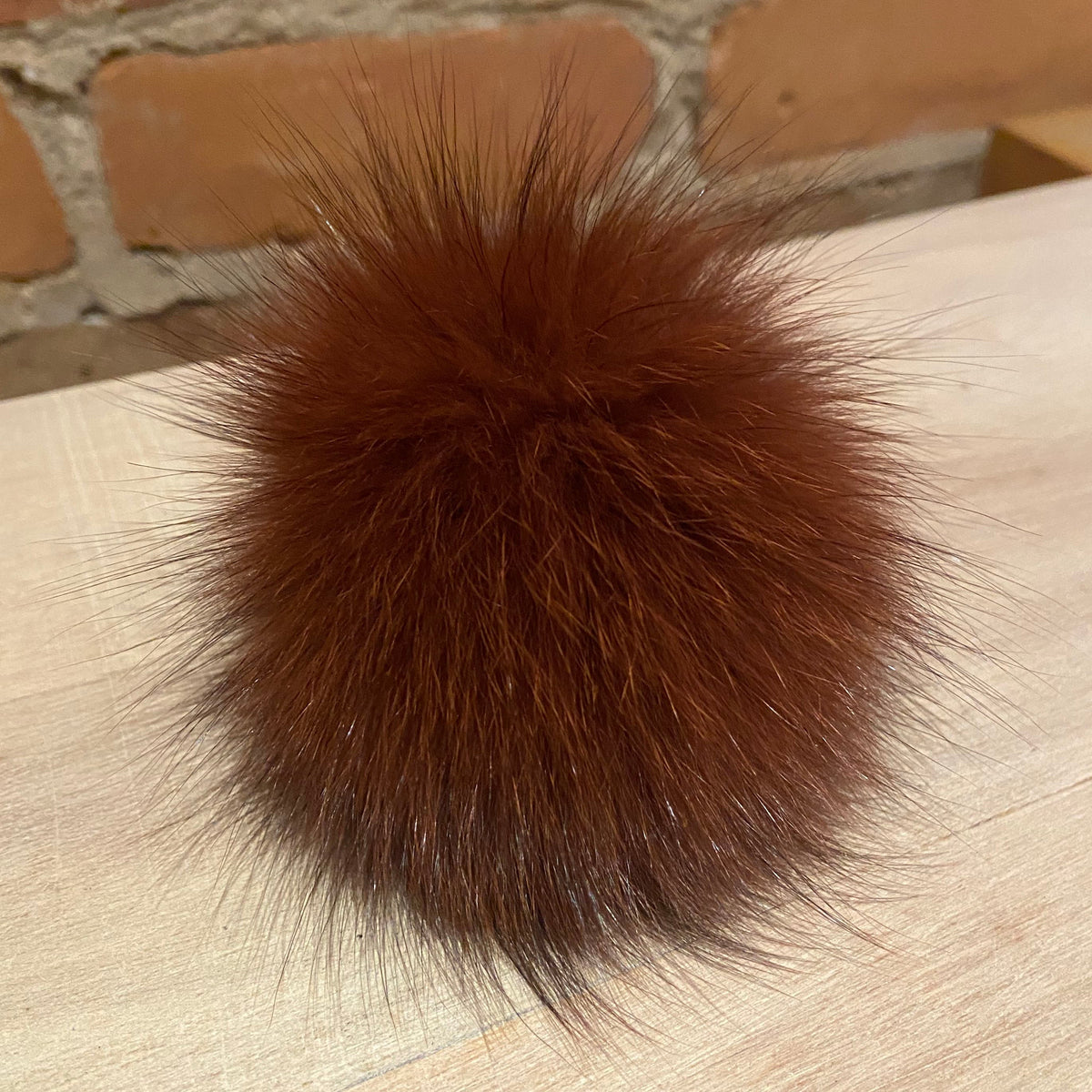 A fluffy, round brown fox fur pom-pom ball resting on a light-colored wooden tabletop with a brick wall in the background.