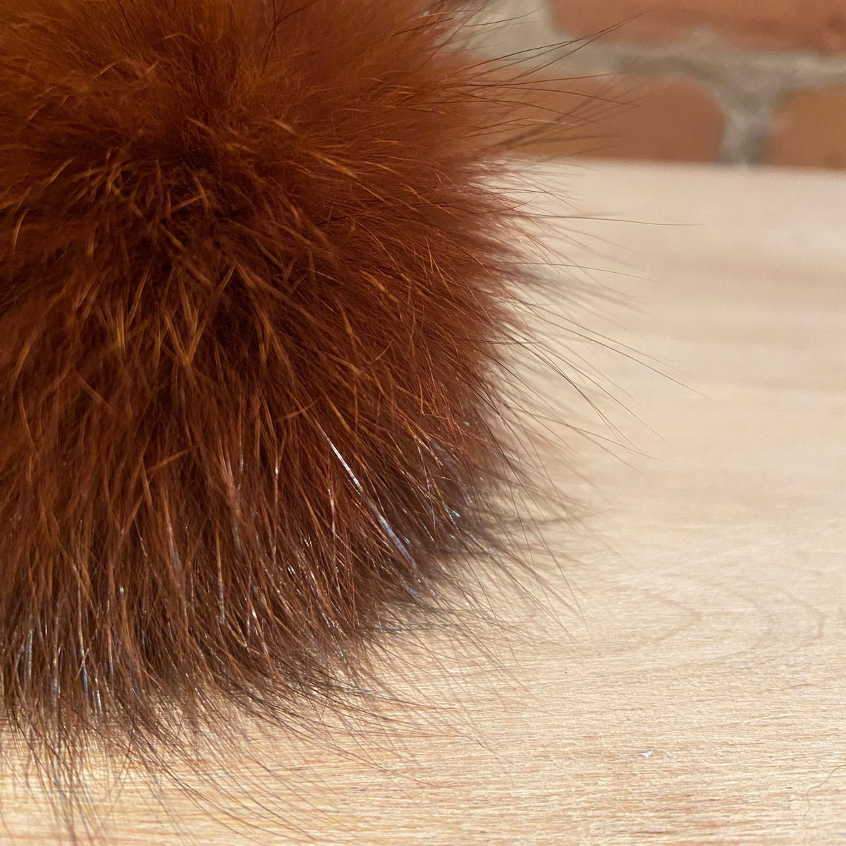A close-up of a fluffy reddish-brown fox fur pom-pom resting on a light wood surface with a blurred brick wall background.