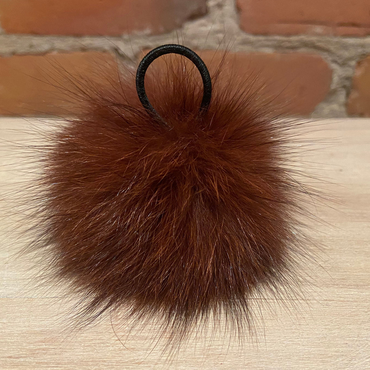 A fluffy brown fox fur pom-pom keychain or bag charm with a black elastic loop, resting on a light wood surface with a brick wall background.