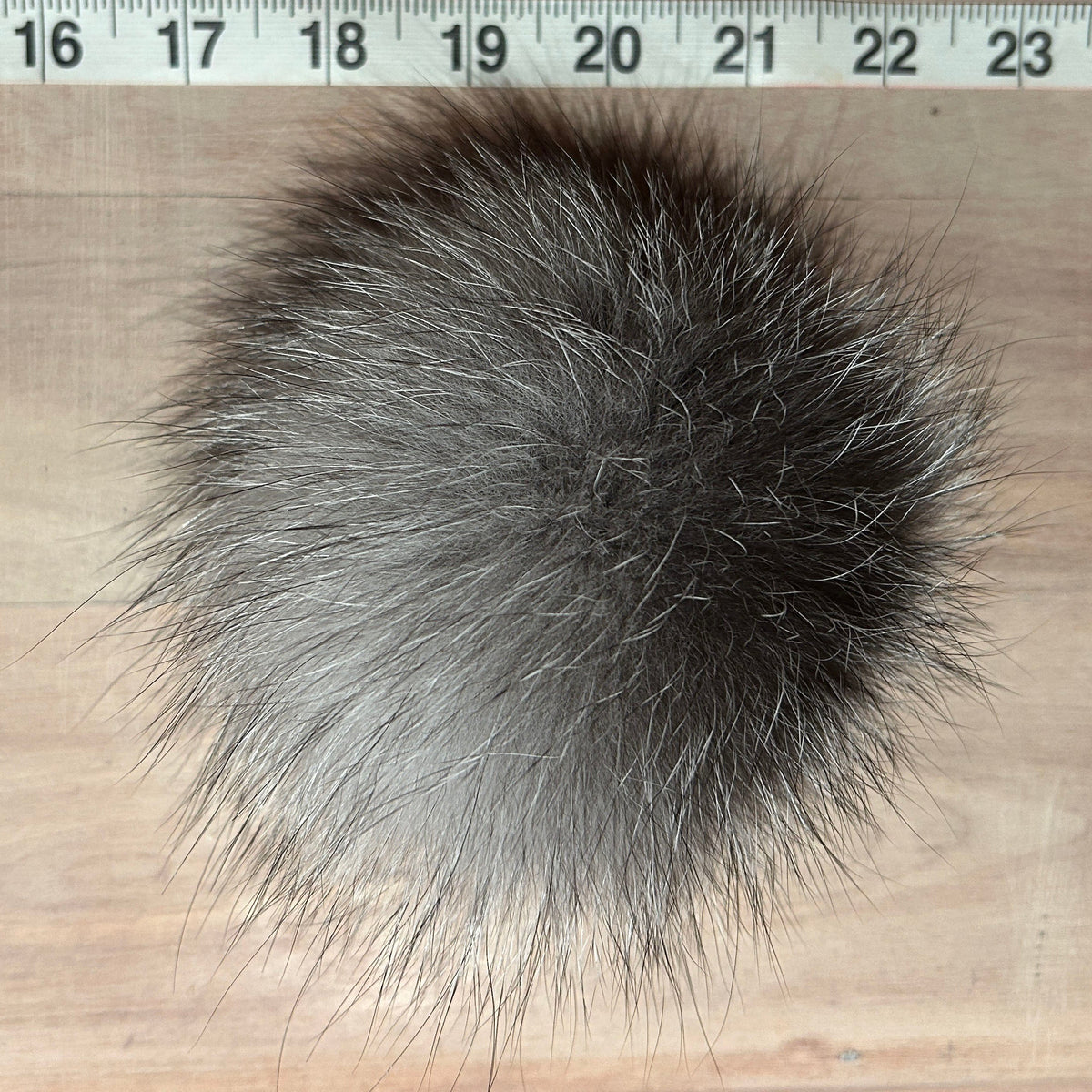 A round, fluffy fox fur pom-pom in a natural gray and black animal fur color, shown on a light wood surface with a measuring tape ruler at the top for scale.