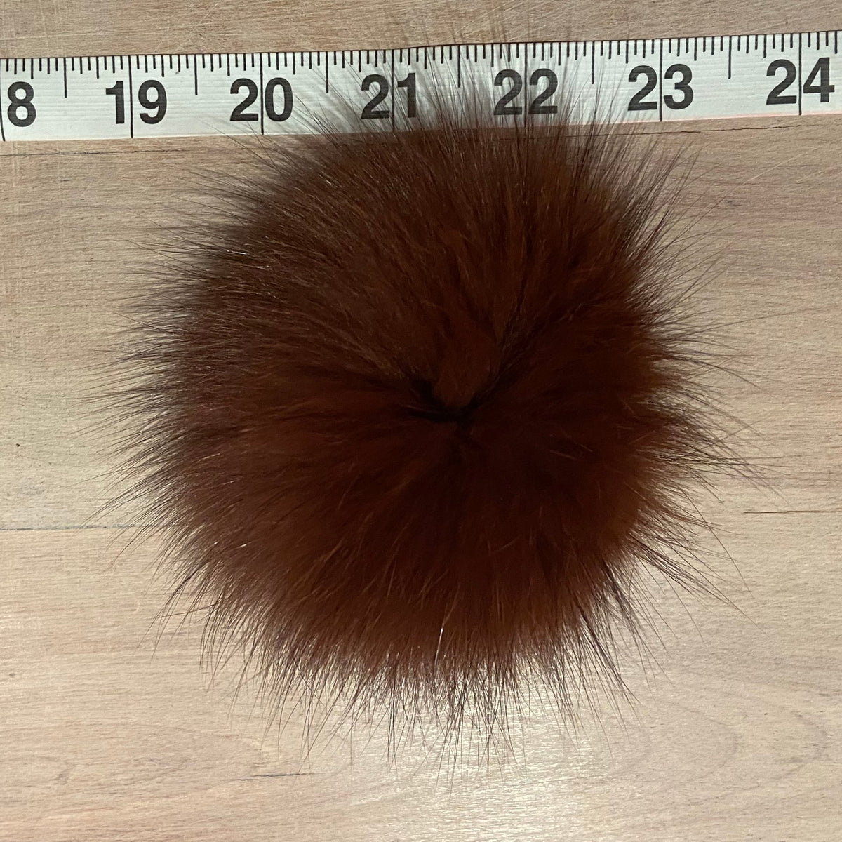 A round brown fox fur pom-pom shown on a light wood surface with a white measuring tape ruler at the top for scale.