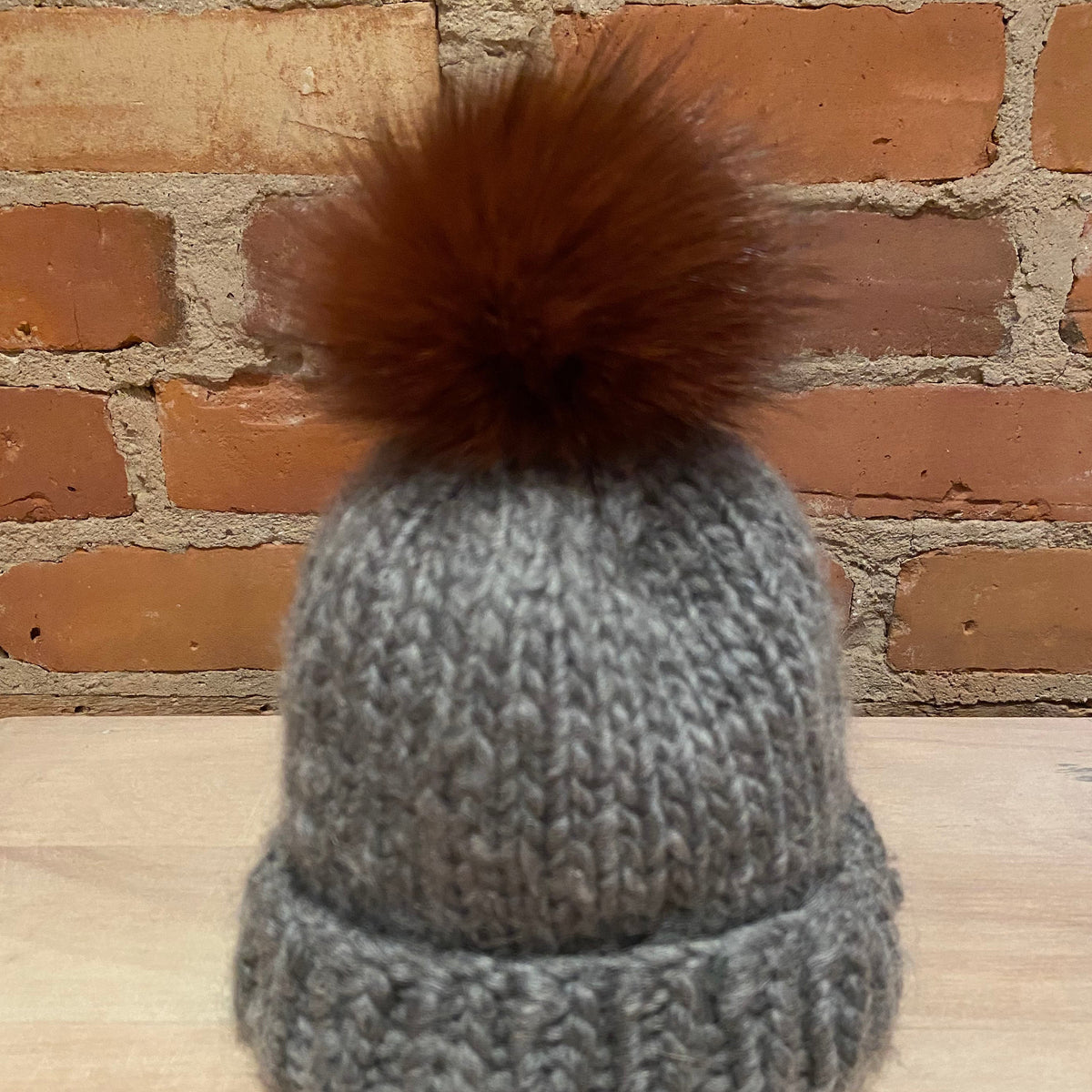 A small  gray chunky knit beanie hat with a small cinnamon fox fur pom-pom, displayed on a light wood surface against a brick wall background.