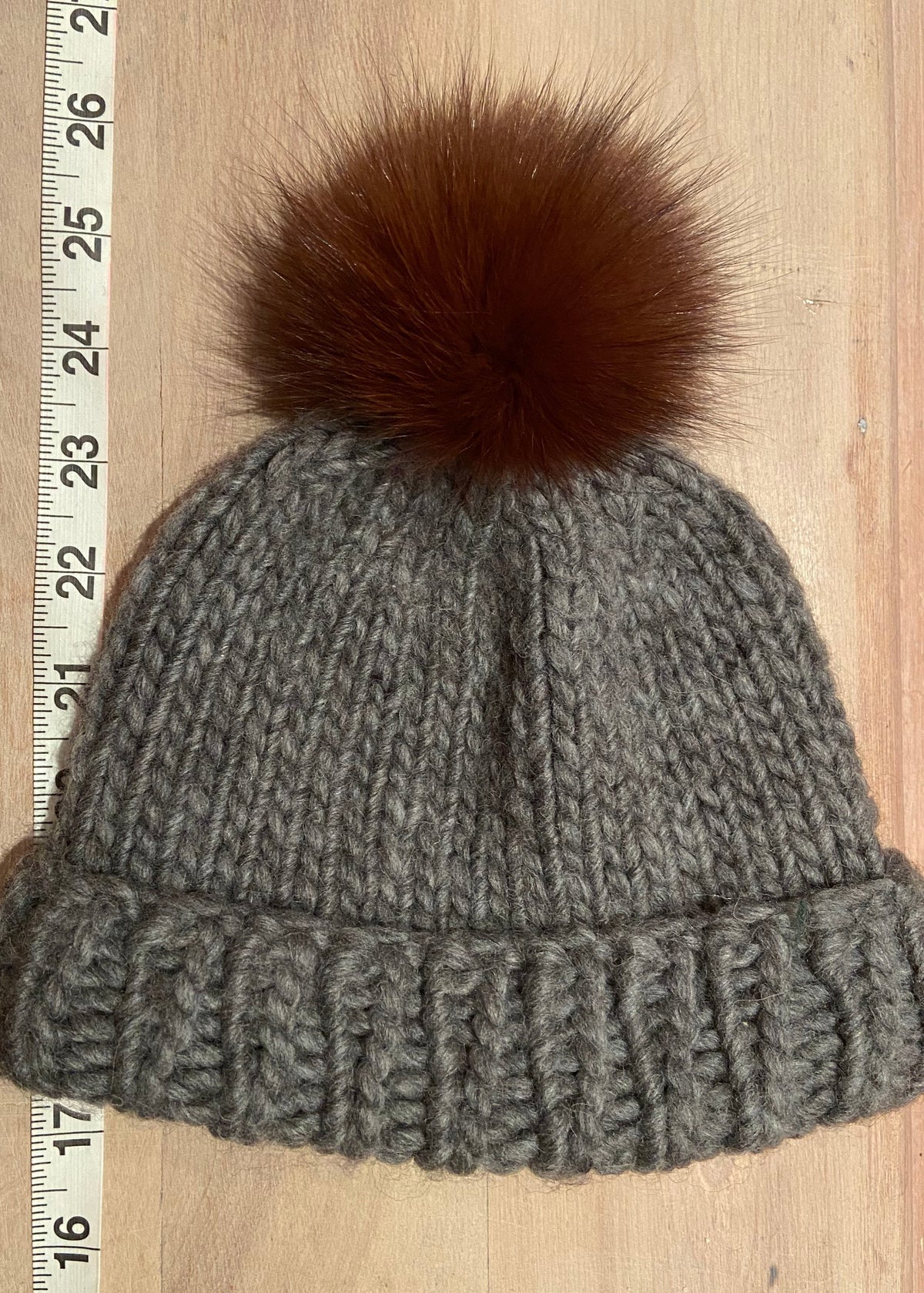 A small gray knit beanie with a small brown fox -fur pom-pom, laid flat on a light wood surface next to a measuring tape.