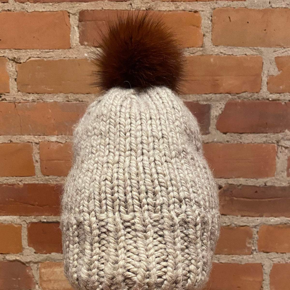 A chunky knit beanie hat in off-white with a brown fox-fur pom-pom, displayed on a mannequin head with red lips in front of a brick wall background.