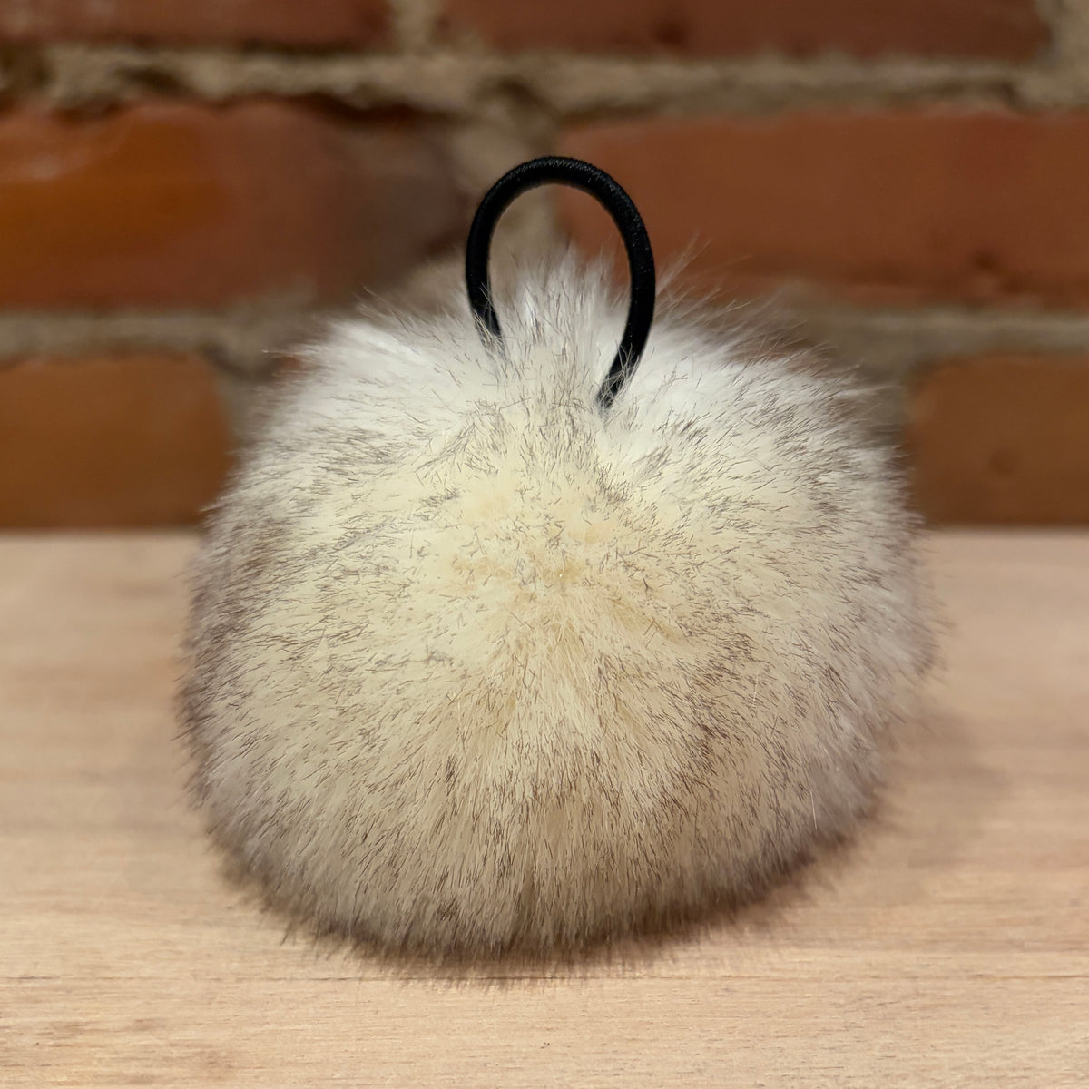 A fluffy faux-fur pom-pom keychain or bag charm in cream and light brown tones, featuring a black elastic loop at the top, resting on a light wood surface with a brick wall background.