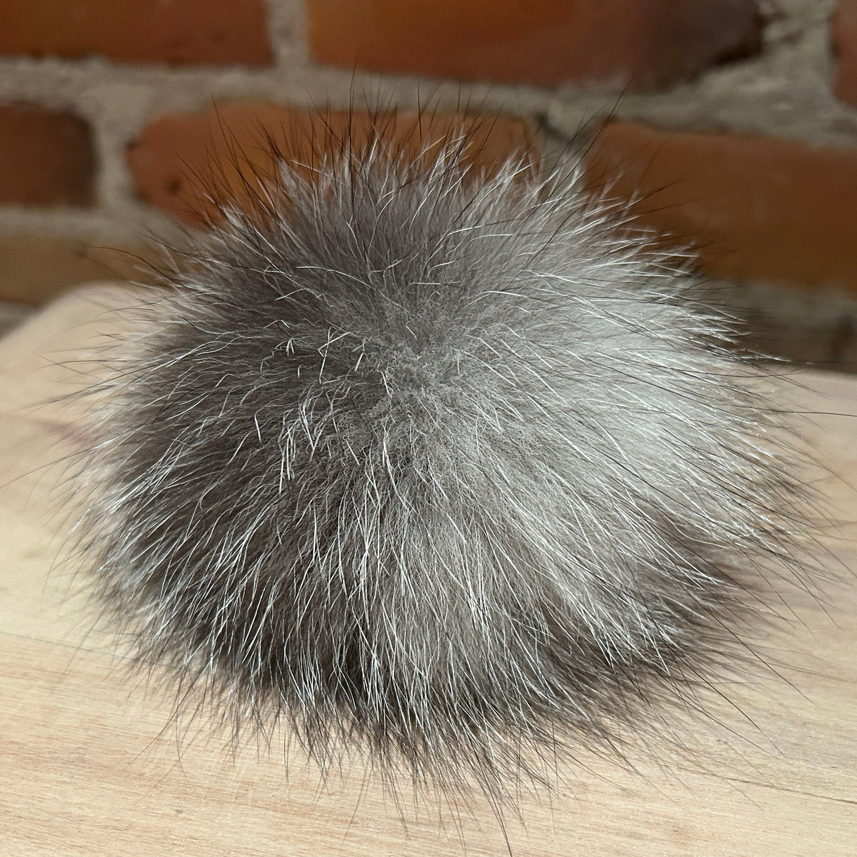 A fluffy pom-pom ball made of gray and silver fox fur, resting on a light wood surface with a blurred brick wall background.