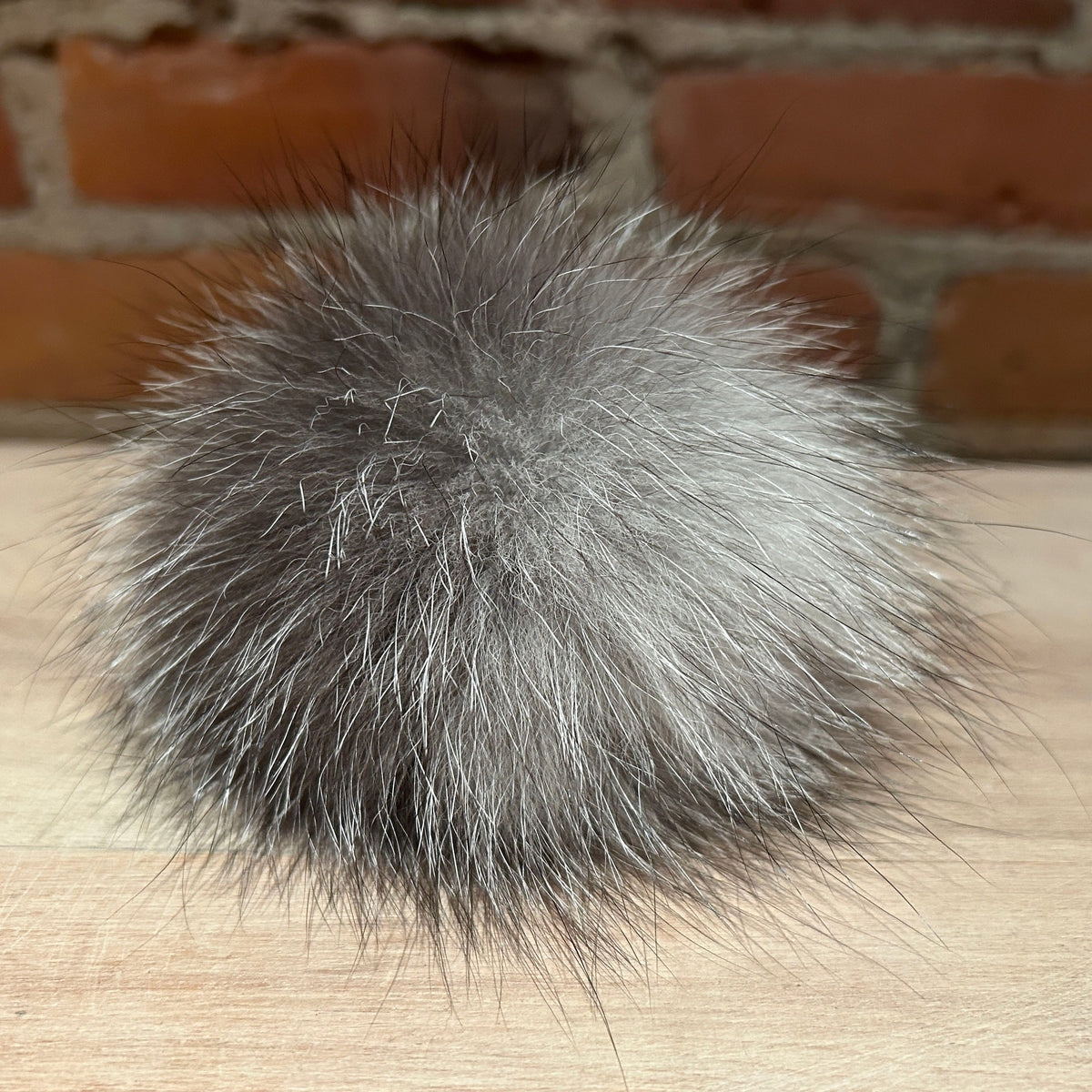 A fluffy gray fox fur pom-pom accessory resting on a light wood surface with a blurred brick wall background.