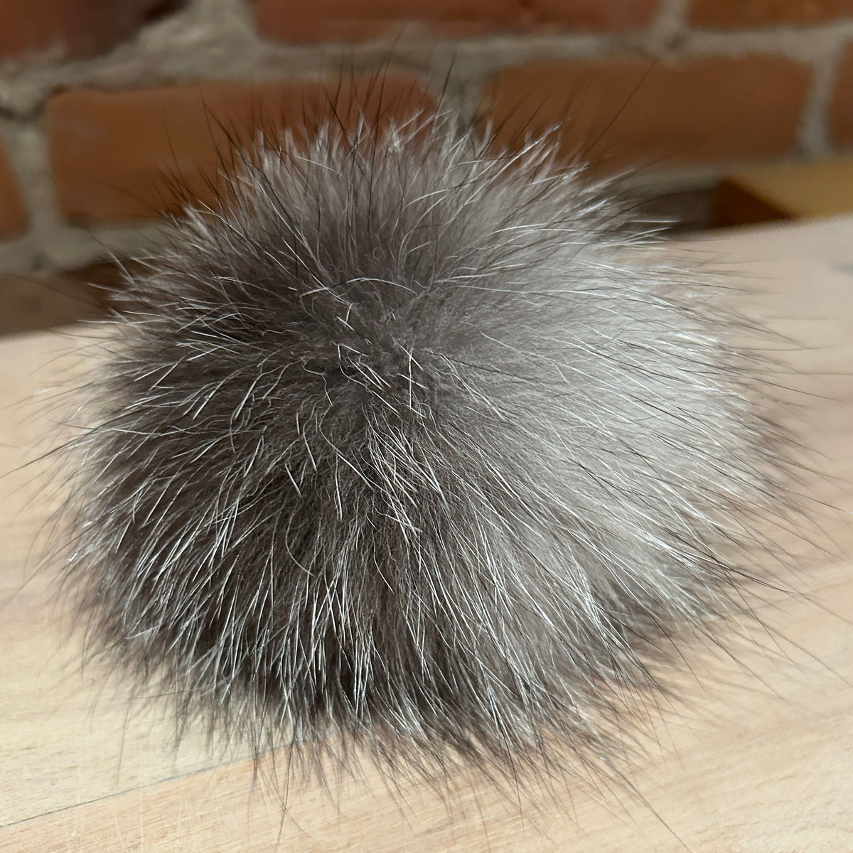 A fluffy pom-pom ball made of gray and silver-toned fox fur or natural fur, resting on a light wood surface with a blurred brick wall in the background.