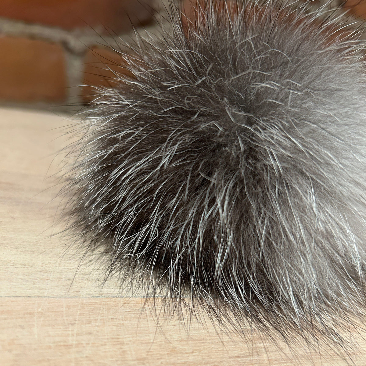 A close-up of a fluffy gray and silver fox fur pom-pom resting on a light wood surface with a blurred brick wall in the background.