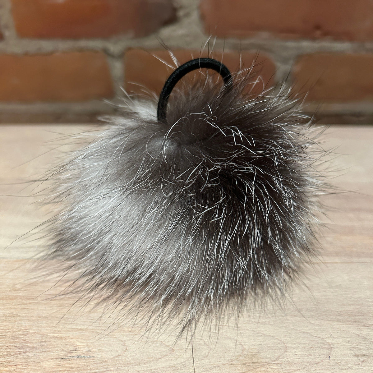 A fluffy fox -fur pom-pom keychain or bag charm in shades of gray, silver, and black, attached to a black elastic band, resting on a light wood surface with a brick wall background.