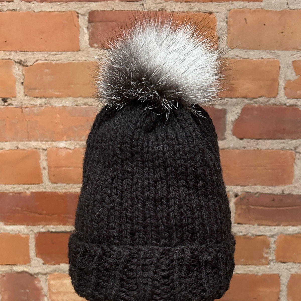A black ribbed knit beanie with a fluffy silver-gray and black pom-pom, displayed on a mannequin head against a red brick wall background.