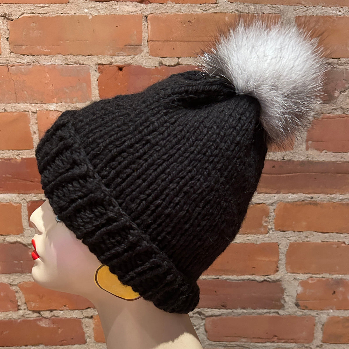 A black ribbed knit beanie hat with a large fluffy white pom-pom, displayed on a mannequin head against a red brick wall background.