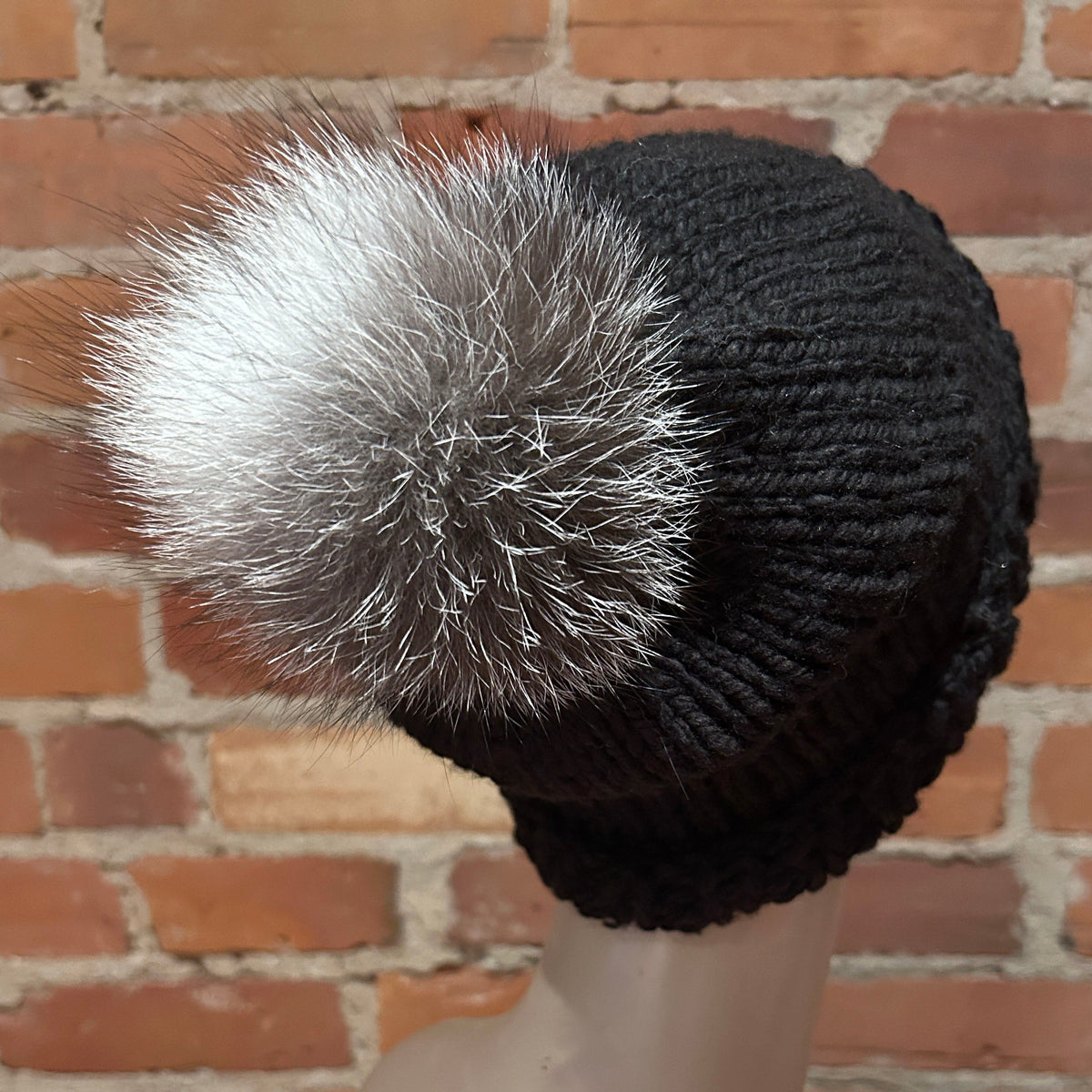 A black ribbed knit beanie hat with a large fluffy silver-gray fox fur pom-pom, displayed on a mannequin head against a red brick wall background.