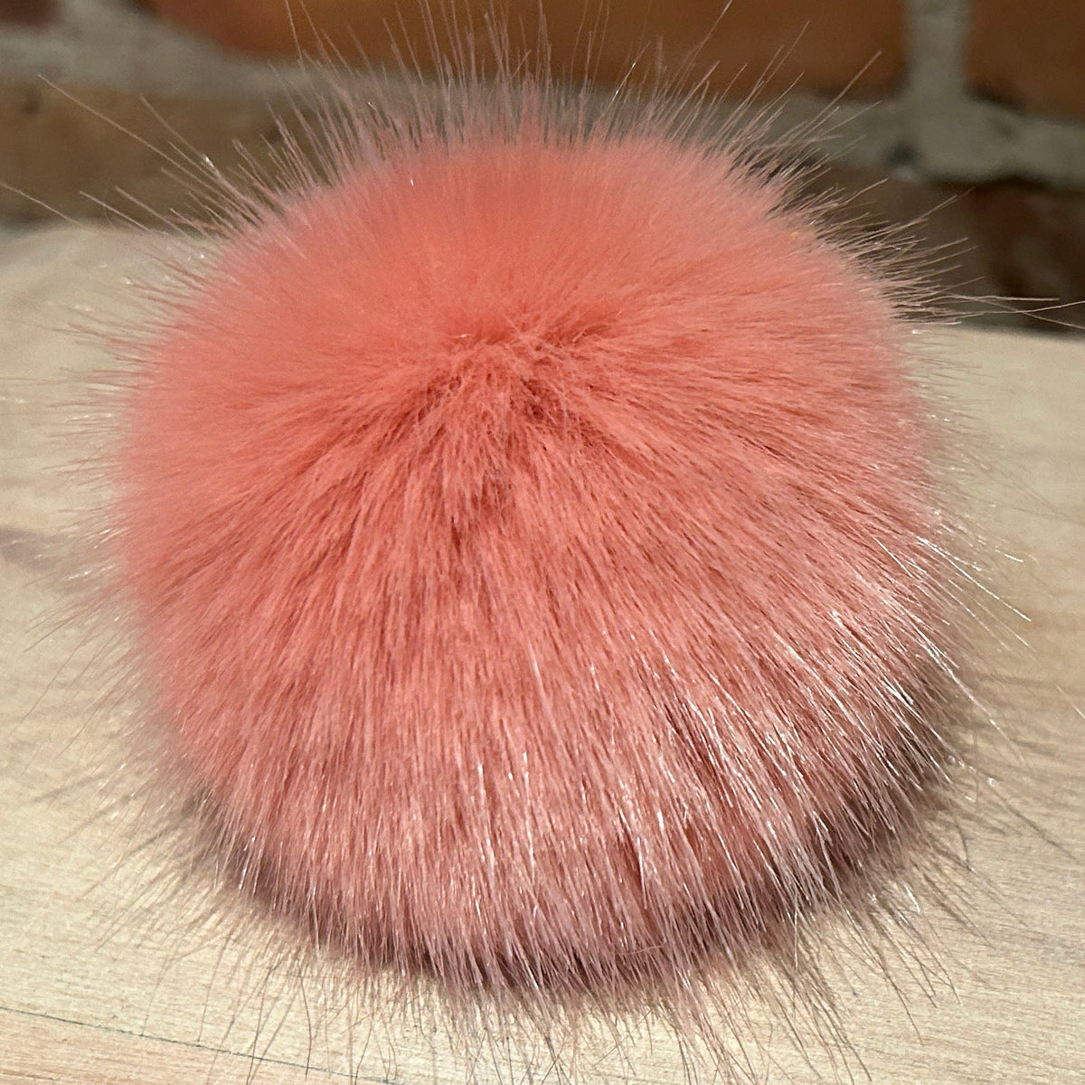 A fluffy, dusty pink faux fur pom-pom accessory resting on a light-colored wooden surface with a brick wall in the background.