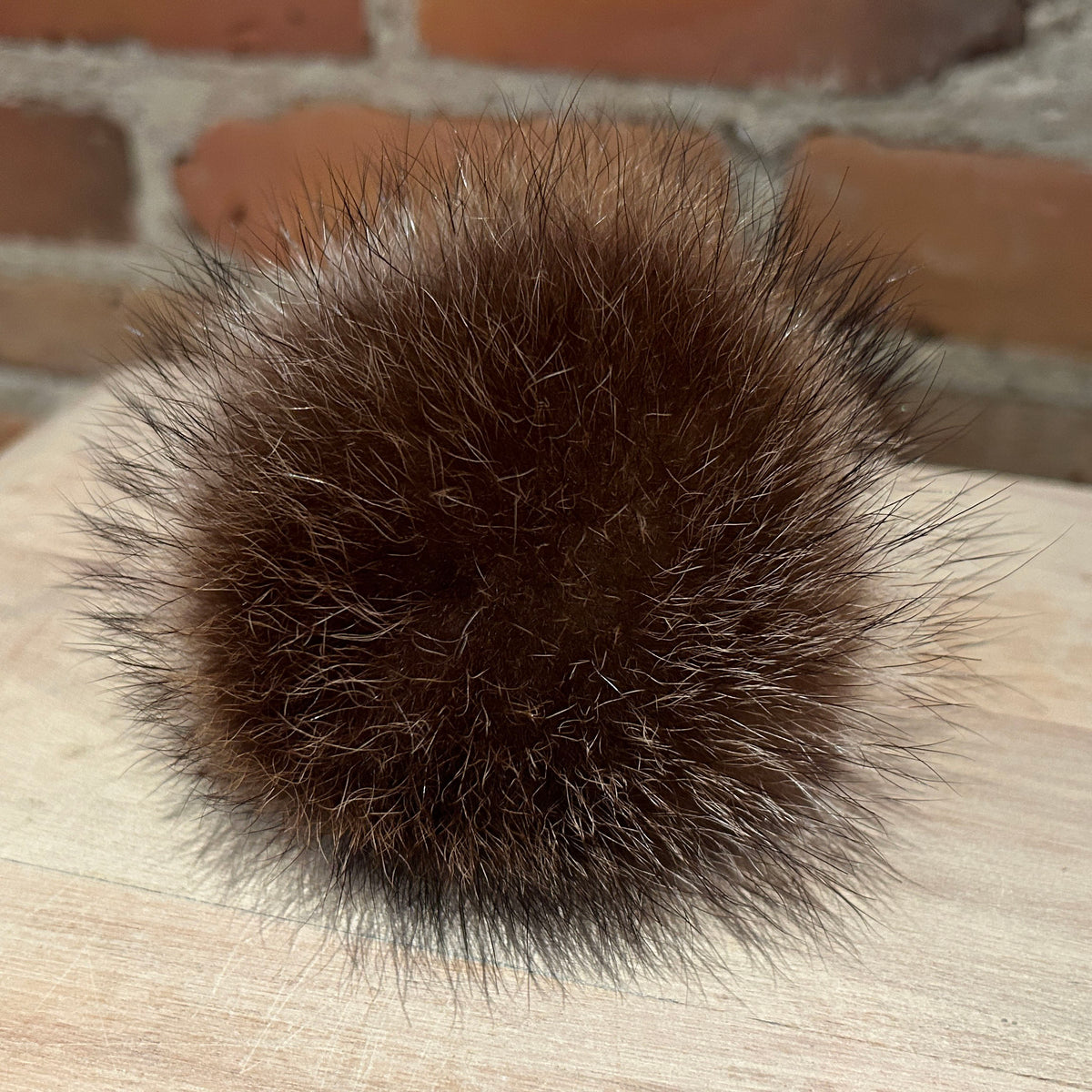 A round, fluffy pom-pom ball made of dark brown fur, resting on a light-colored wooden surface with a blurred brick wall in the background.