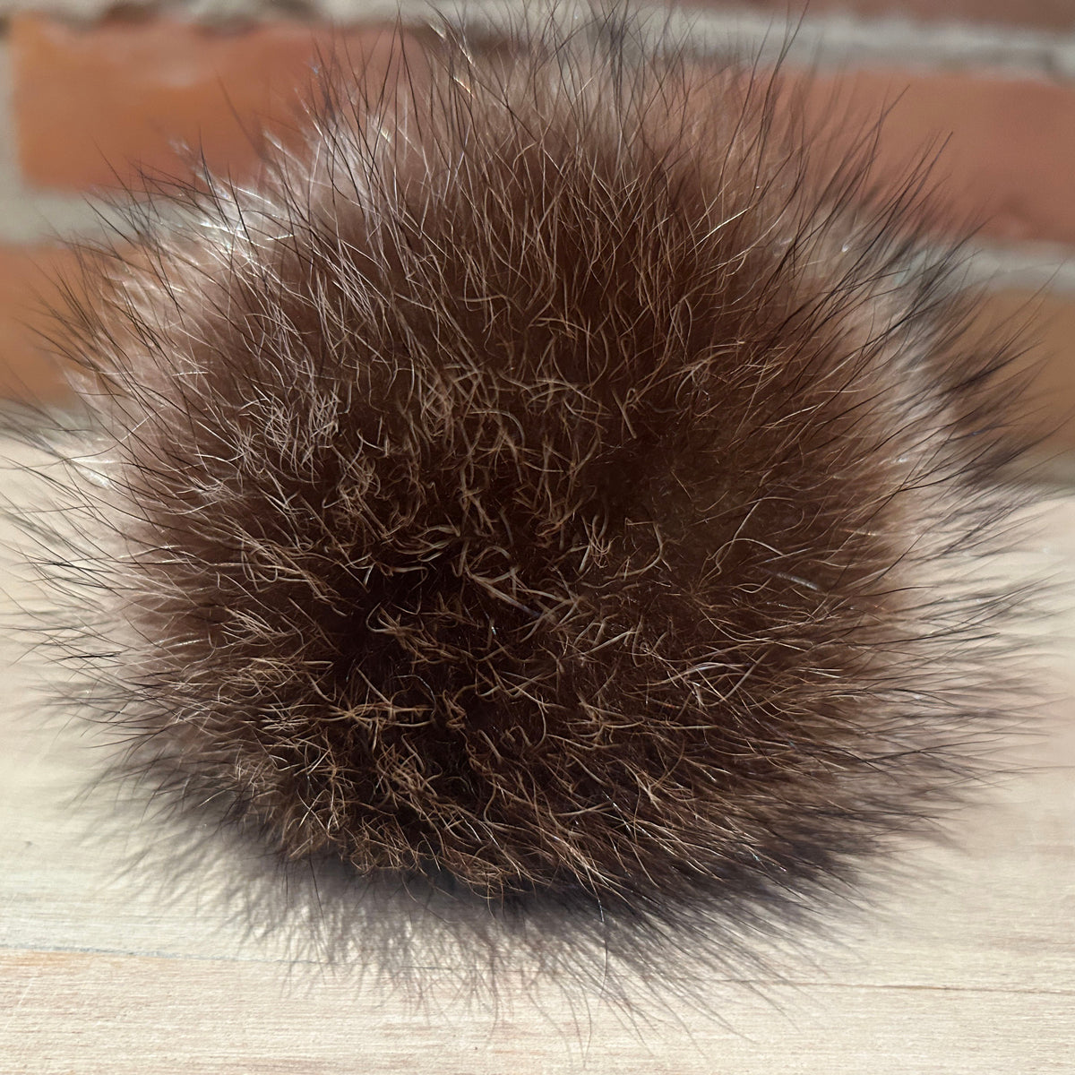 A close-up of a dark brown fluffy pom-pom, likely made from fur or faux fur, resting on a light wood surface with a blurred brick wall in the background.
