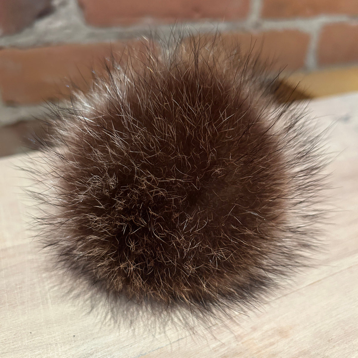 A round, fluffy pom-pom ball made of dark brown fur with lighter tan and cream tips, resting on a light wood surface with a blurred brick wall in the background.