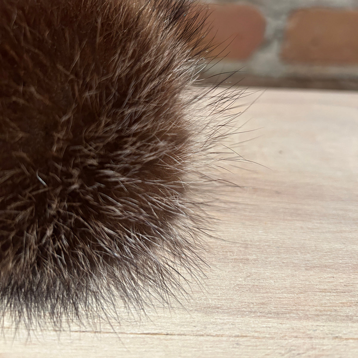 A close-up of a dark brown raccoon fur pom-pom resting on a light wood surface with a blurred brick wall background.