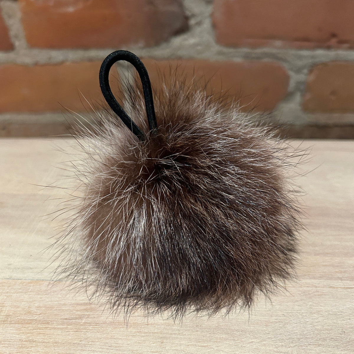 A brown and black raccoon fur pom-pom keychain or bag charm with a black elastic loop, resting on a light wood surface with a brick wall background.