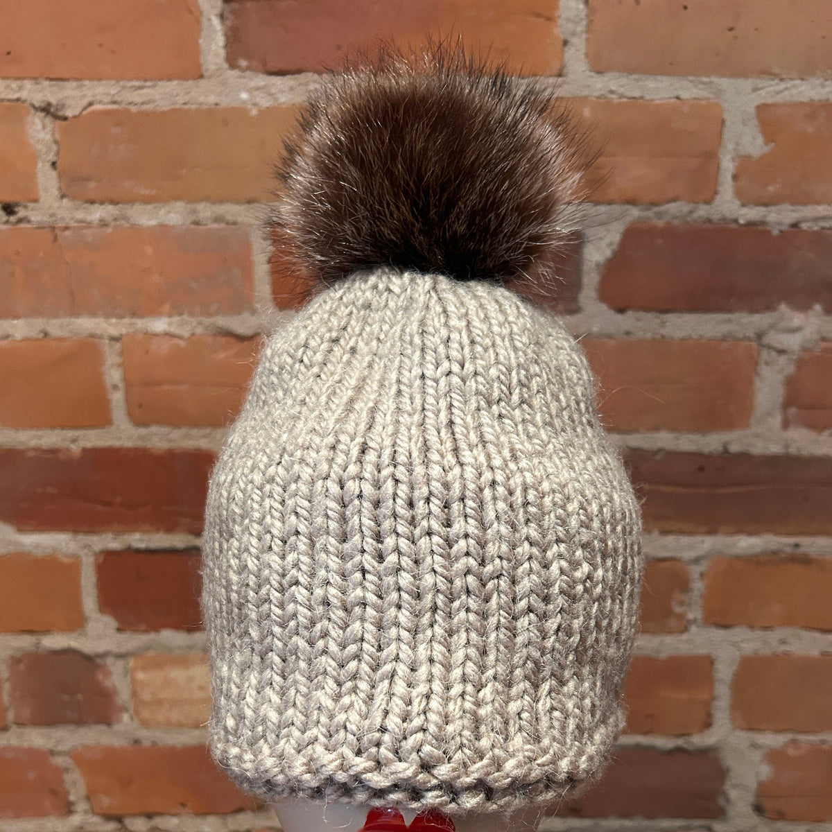 A chunky knit beanie hat in a light oatmeal gray color, topped with a large dark brown raccoon fur pom-pom. The hat is displayed on a white mannequin head with red lipstick, set against a rustic red-brown brick wall background.