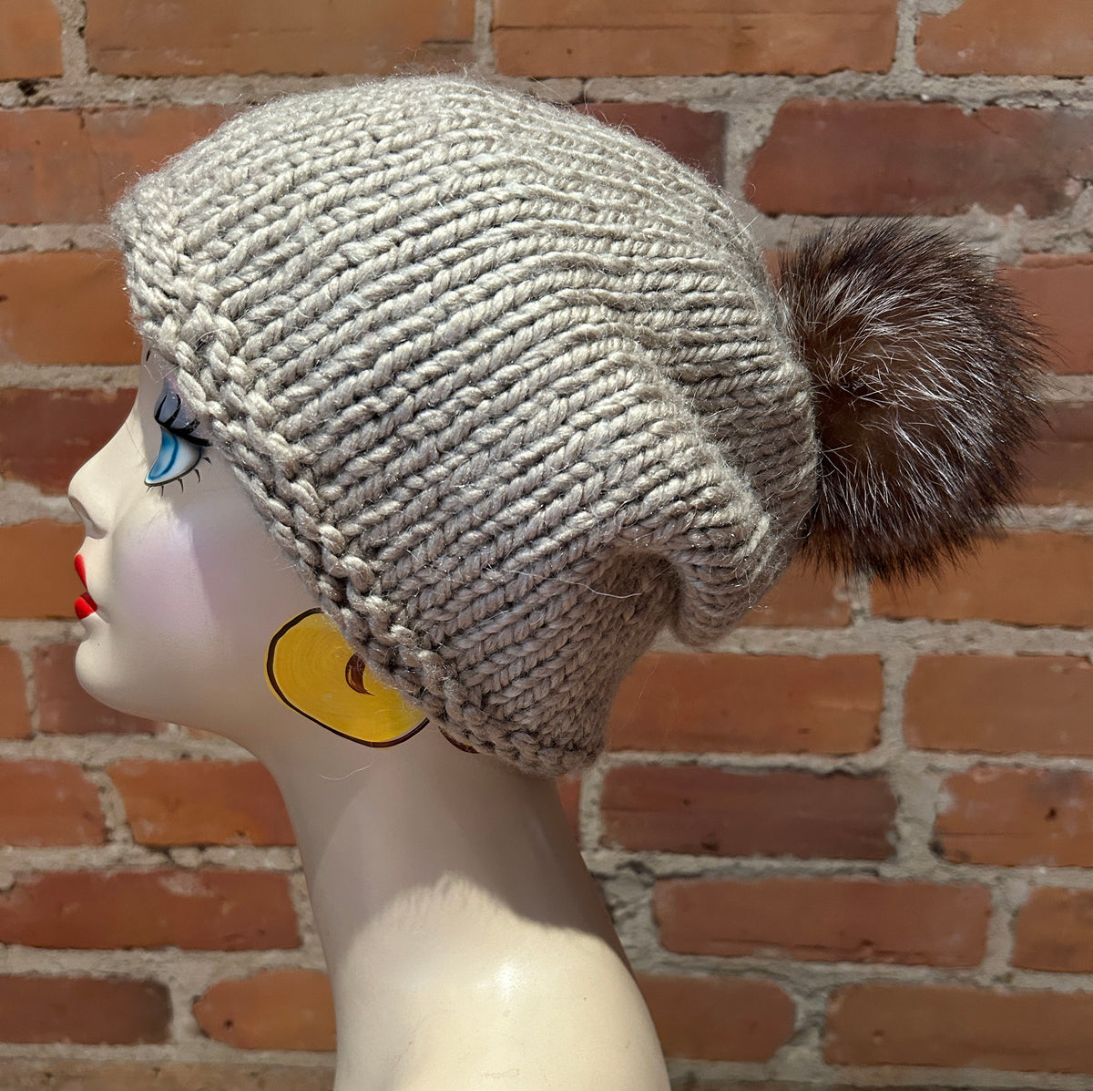 A light gray chunky knit beanie with a large brown and white raccoon fur pom-pom, displayed on a mannequin head against a red brick wall background.