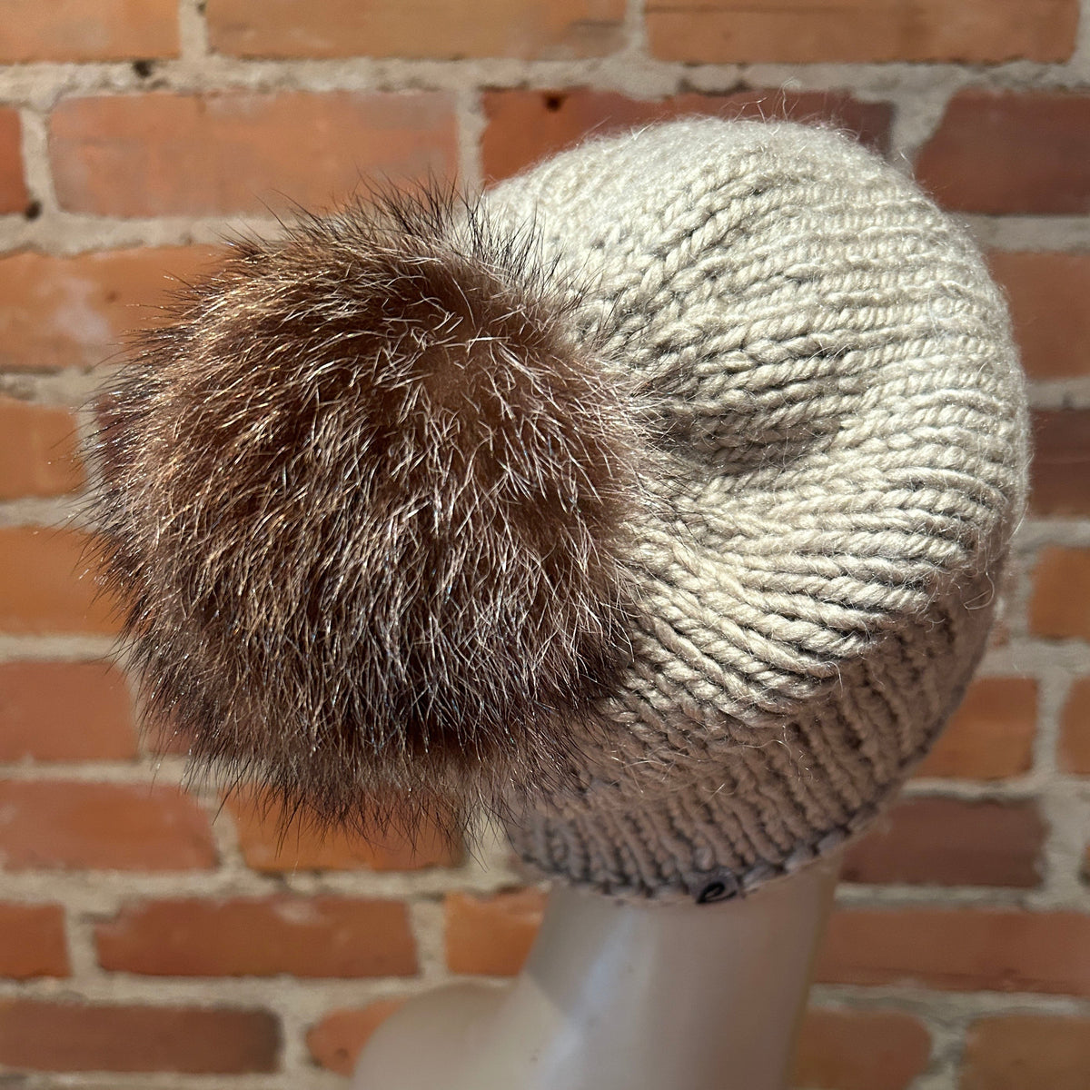 A cream-colored chunky knit beanie hat with a large brown and beige raccoon fur pom-pom, displayed on a mannequin head against a brick wall background.