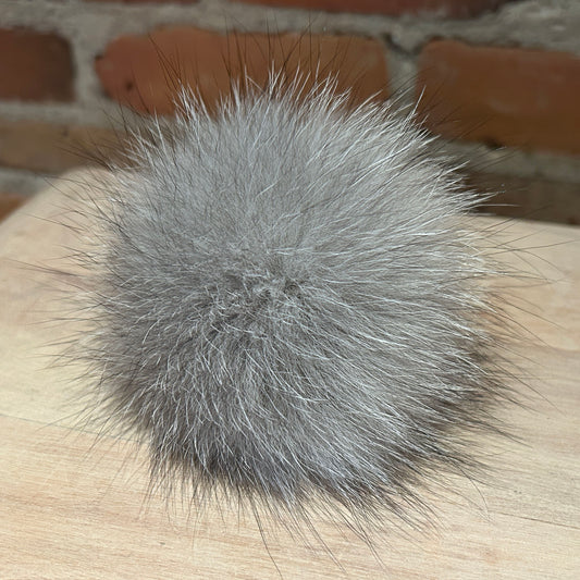 A fluffy gray fox fur pom-pom accessory resting on a light wood surface with a blurred brick wall background.