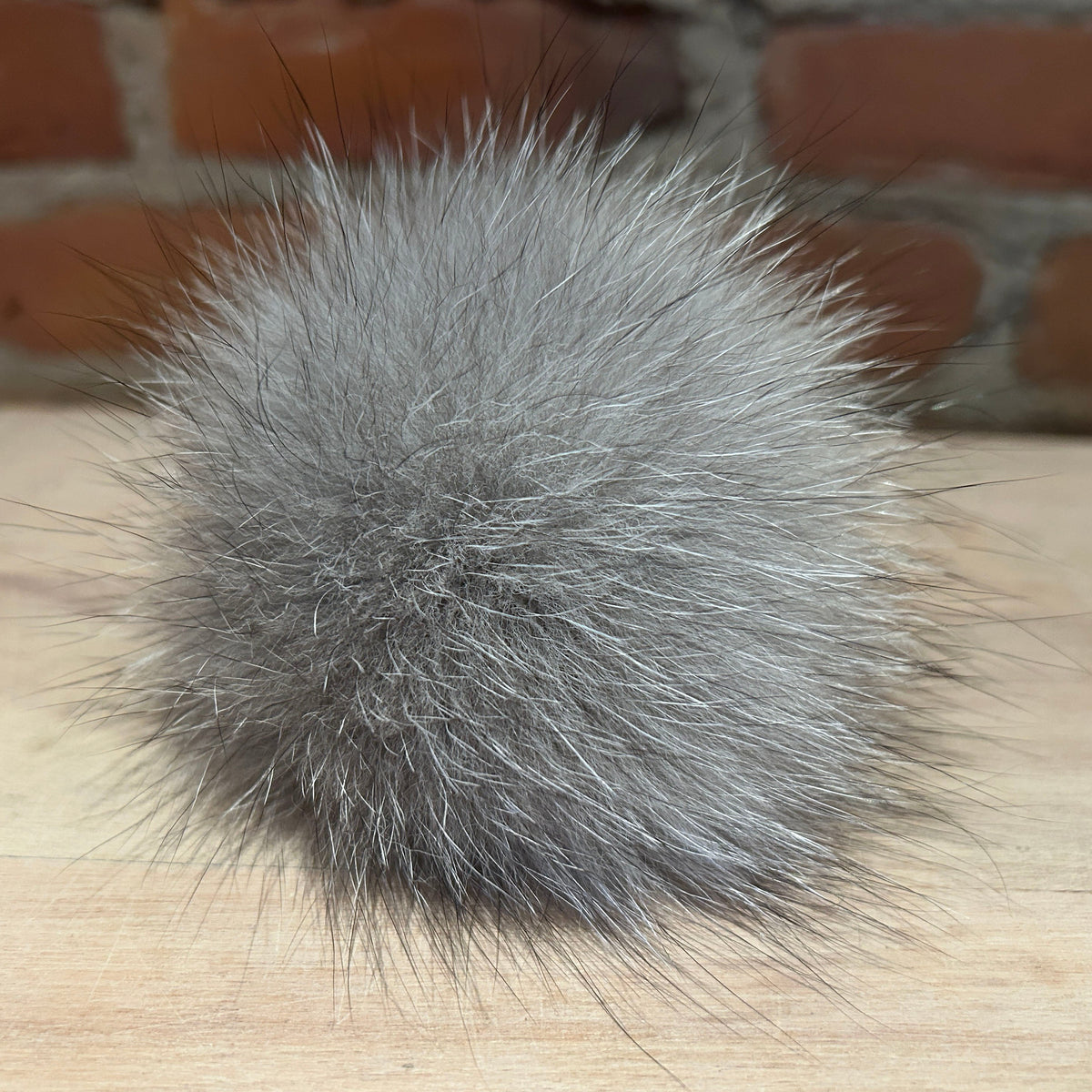 A fluffy gray fox fur pom-pom ball resting on a light wood surface with a blurred brick wall background.
